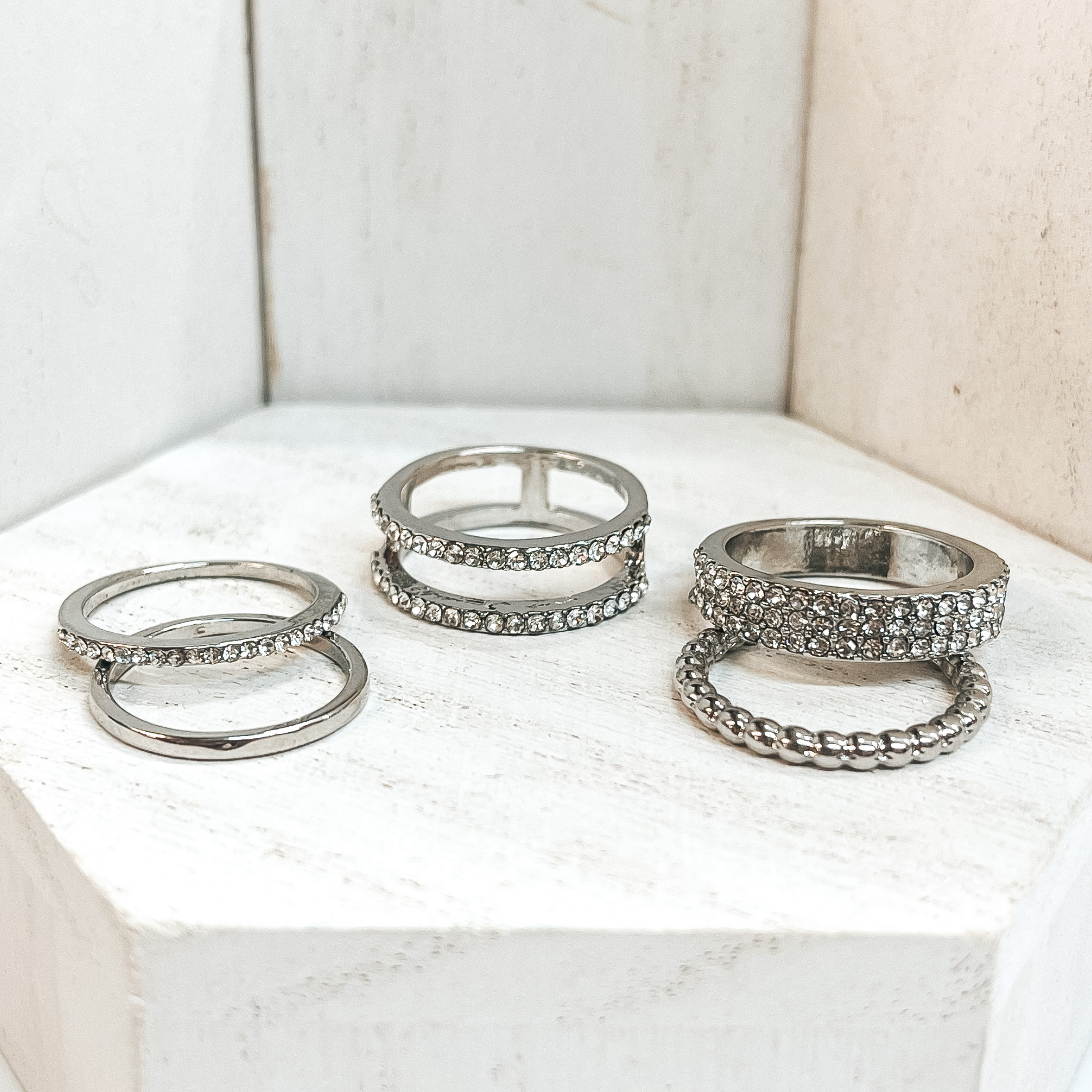 This is a set of five silver rings. All of them but  two rings have crystals in different varieties on  them. One of the rings is bubbled textured and the  other is a solid band. These rings are pictured on a white block and  white background.