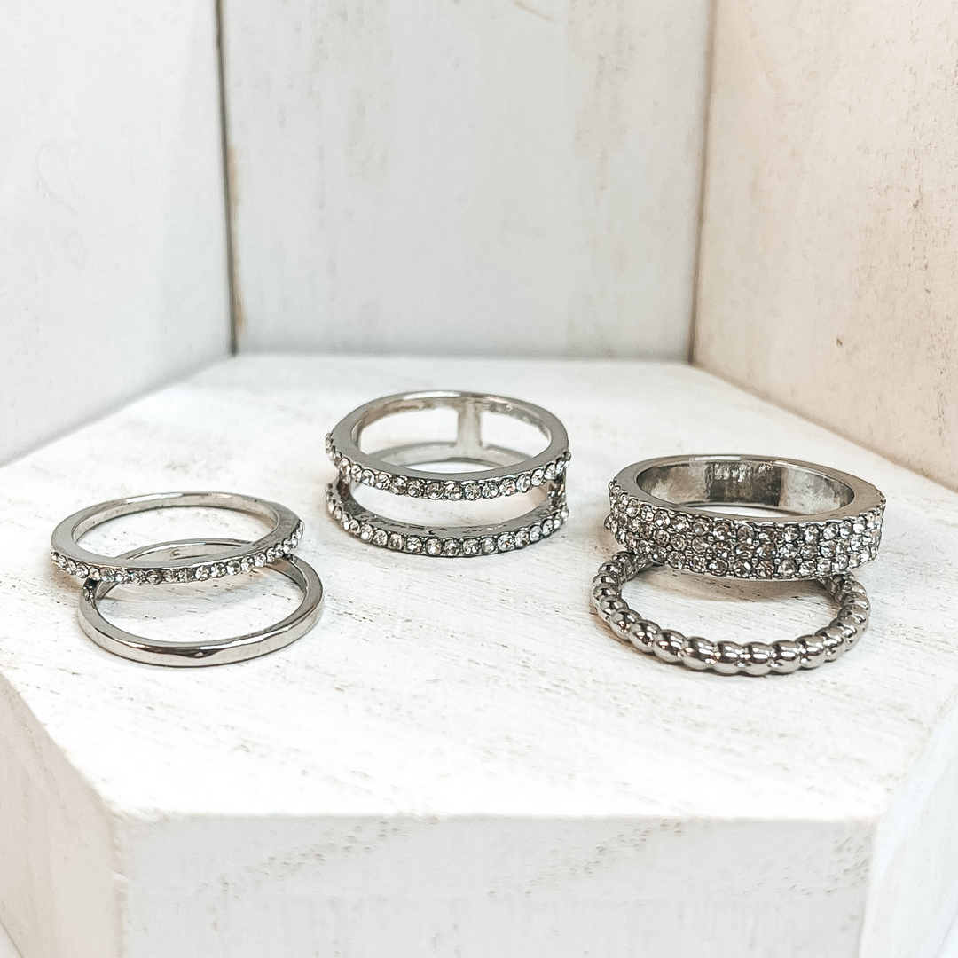 This is a set of five silver rings. All of them but  two rings have crystals in different varieties on  them. One of the rings is bubbled textured and the  other is a solid band. These rings are pictured on a white block and  white background.