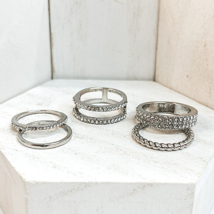 This is a set of five silver rings. All of them but  two rings have crystals in different varieties on  them. One of the rings is bubbled textured and the  other is a solid band. These rings are pictured on a white block and  white background.