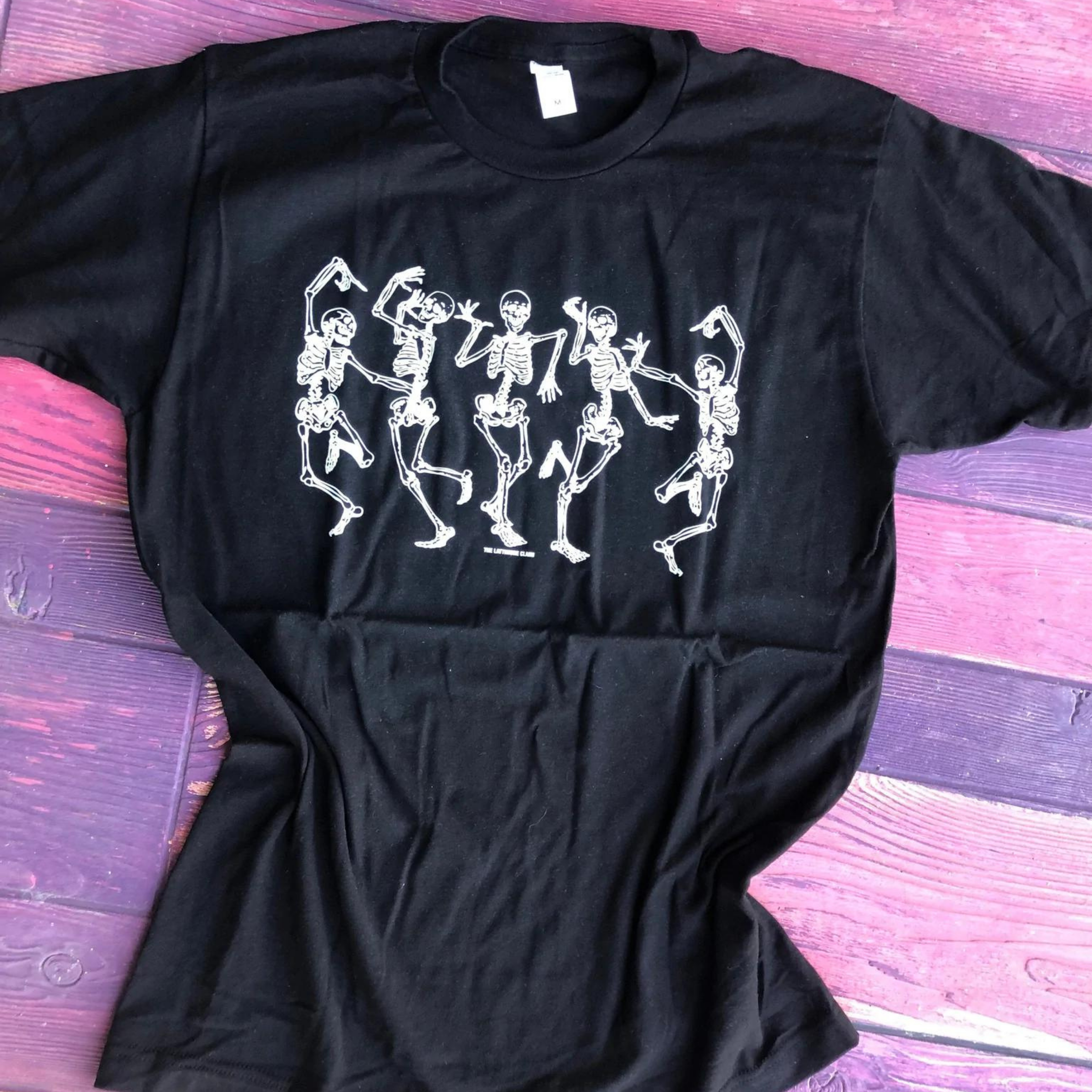 Black short sleeve graphic tee with five dancing skeletons. The background of this picture is purple wood.