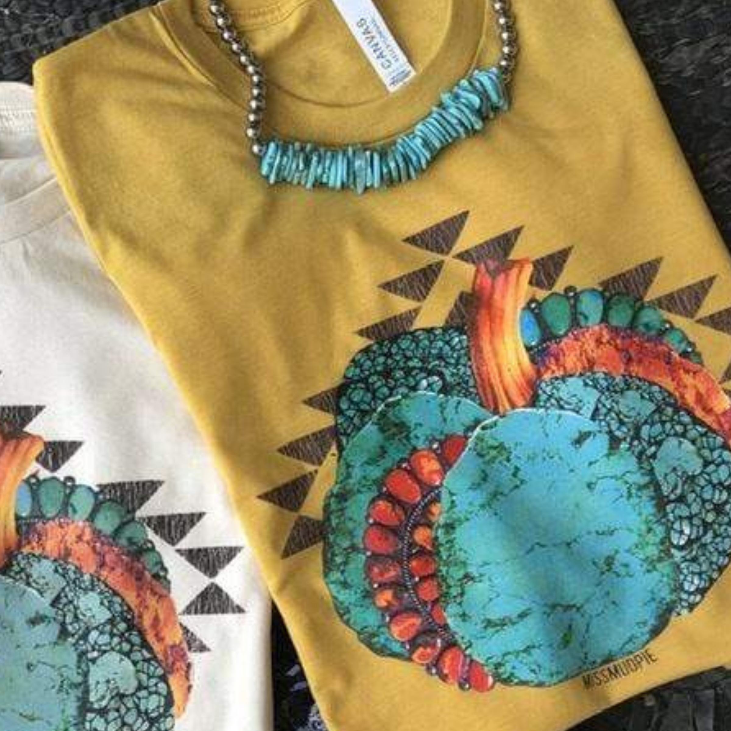 This is a mustard yellow graphic tee with a turquoise pumpkin. There is a red accent piece of jewelry in the side of the pumpkin. There are multiple many triangles surrounding the pumpkin. This top is paired with a large chunky turquoise necklace.   