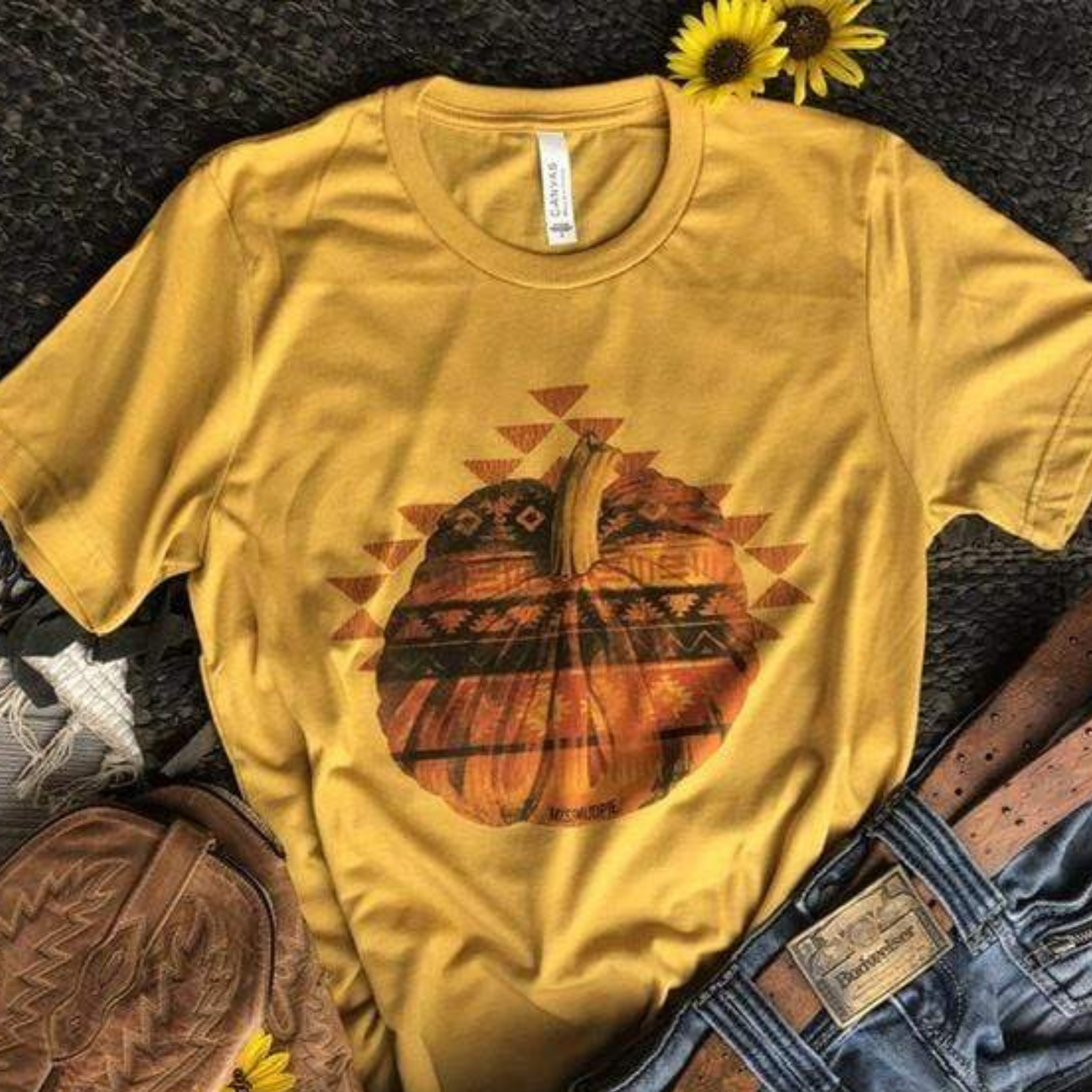 This is a mustard yellow tee with an Aztec designed pumpkin. Surrounding the pumpkin is several many triangles. This top is pictured with some blue jeans and brown cowgirl boots along with two little sunflowers. 