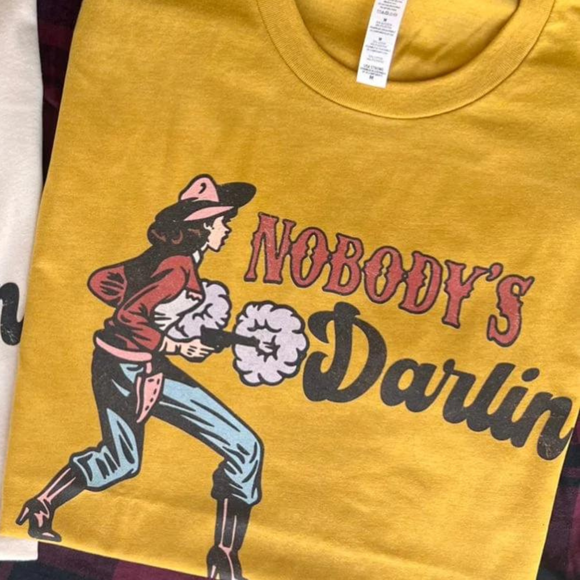 This mustard yellow graphic tee includes a crew neckline, short sleeves, and cute hand drawn design of a cowgirl facing to the side holding pistols and the words "NOBODY'S DARLIN'" in red western font and black cursive. This tee is shown in this photo as a folded flat lay.