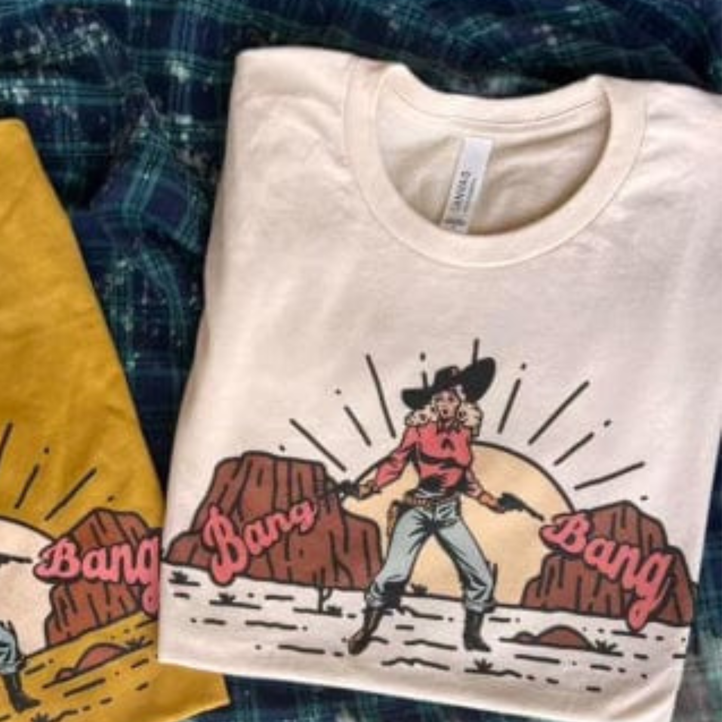 This cream graphic tee includes a crew neckline, short sleeves, and cute hand drawn design of a western scene of a cowgirl in front of mountains and the sun with pistols and the words "Bang Bang" in pink bubble font on either side of her. This tee is shown in this photo as a folded flat lay.