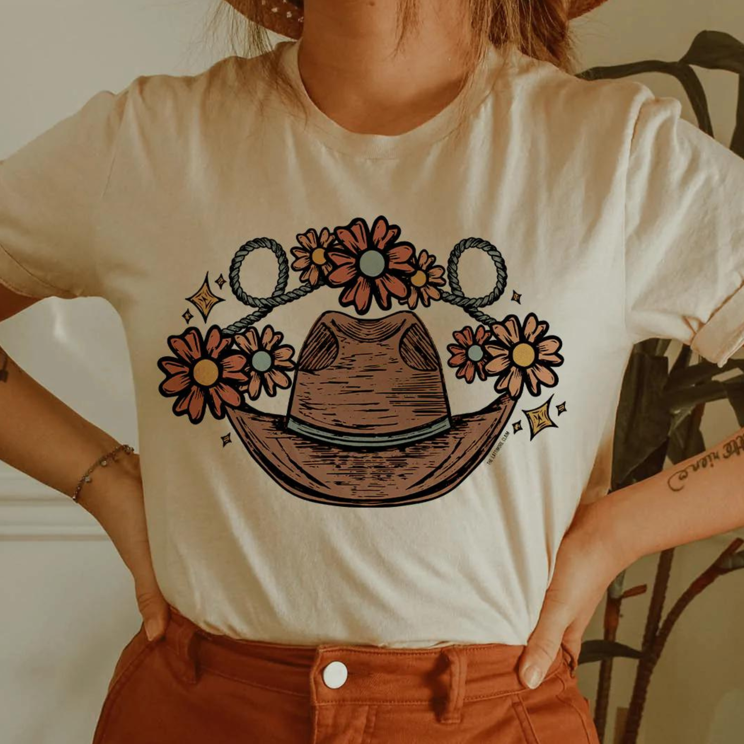 This cream graphic tee includes a crew neckline, short sleeves, and cute hand drawn design of a cowboy hat, rope, stars, and flowers. This tee is shown in this photo styled with a straw hat, silver dainty bracelet, and rust jeans.
