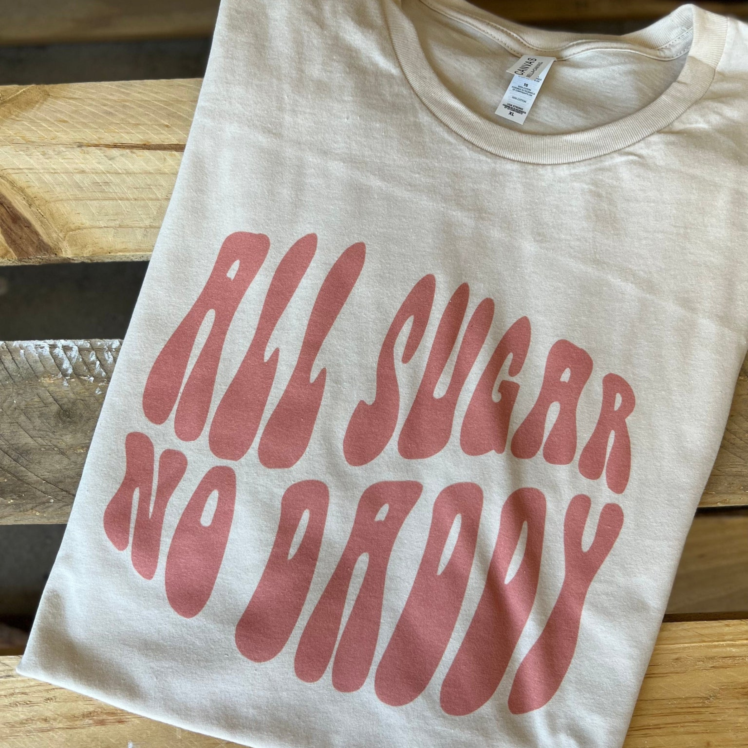This cream Bella + Canvas graphic tee includes a crew neckline, short sleeves, and cute hand drawn design that says "ALL SUGAR NO DADDY" in a pink bubble type of font. This tee is shown in this photo as a folded flat lay.
