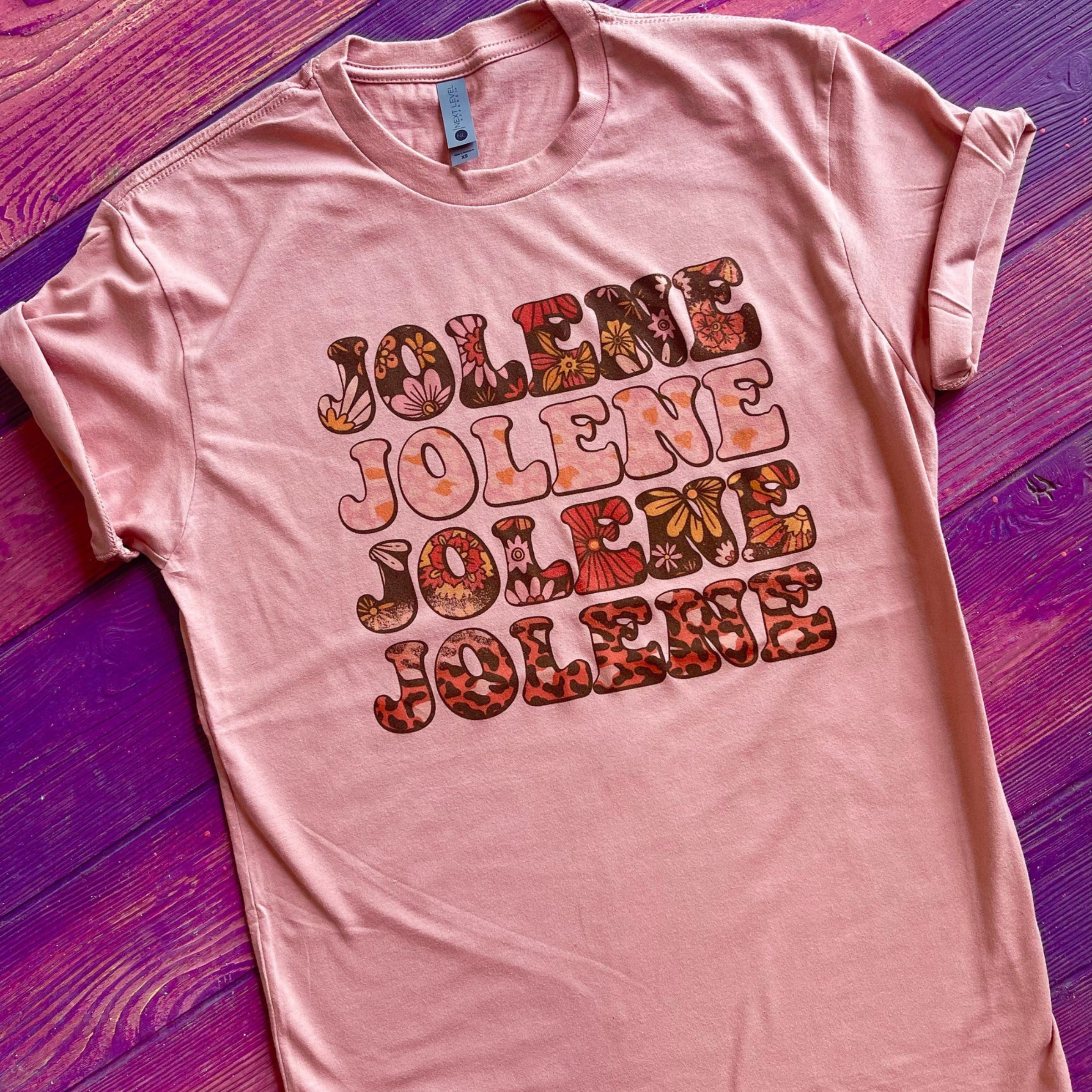 This desert rose pink graphic tee includes a crew neckline, short sleeves, and cute hand drawn design that says "JOLENE" in four rows. Inside of each "JOLENE" is a different print, such as floral or leopard. This tee is shown in this photo as a flat lay with rolled sleeves. 