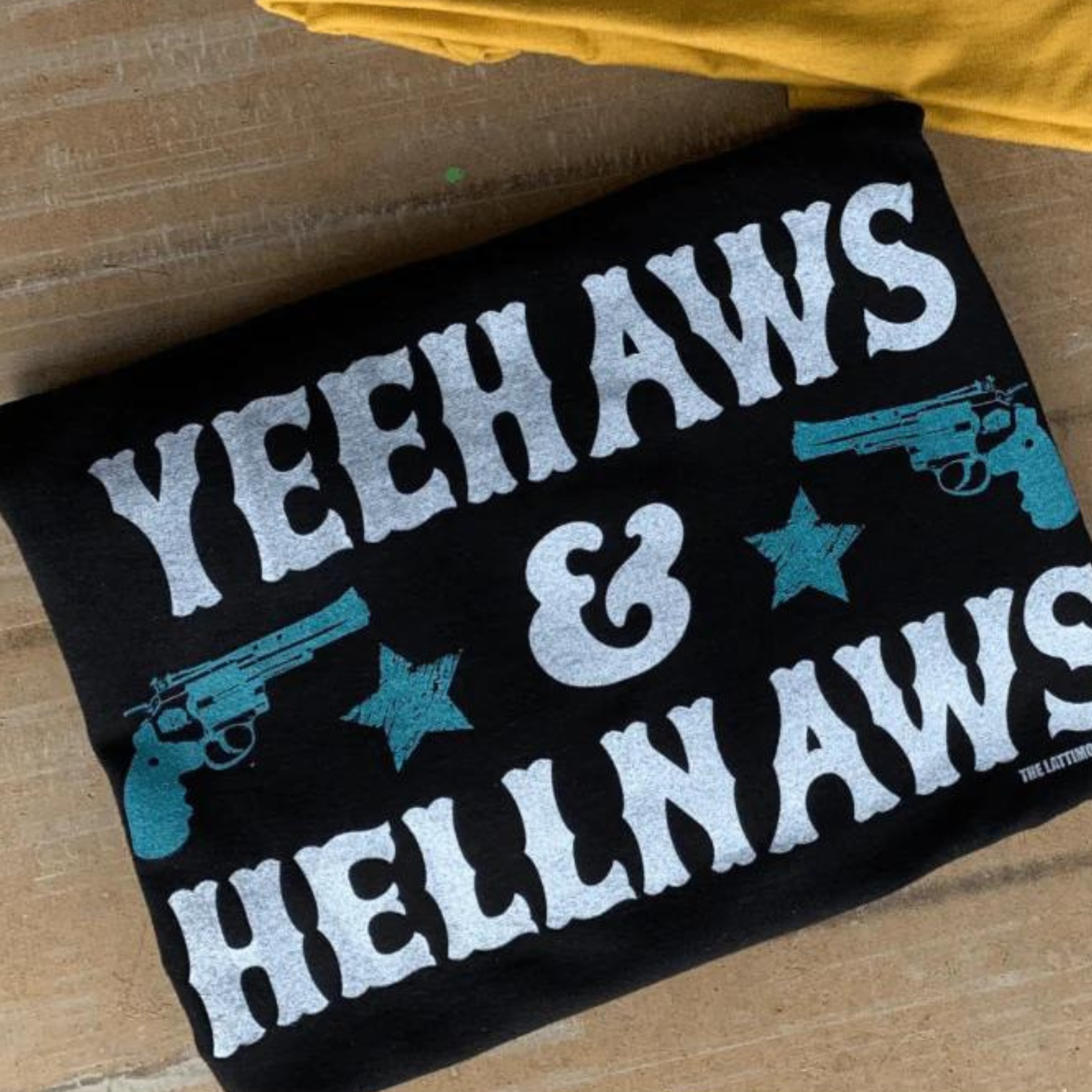 This black graphic tee includes a crew neckline, short sleeves, and cute hand drawn design of two turquoise guns and stars on either side of the & in the saying "YEEHAWS & HELLNAWS" which is stacked. This tee is shown in this photo as a folded flat lay.