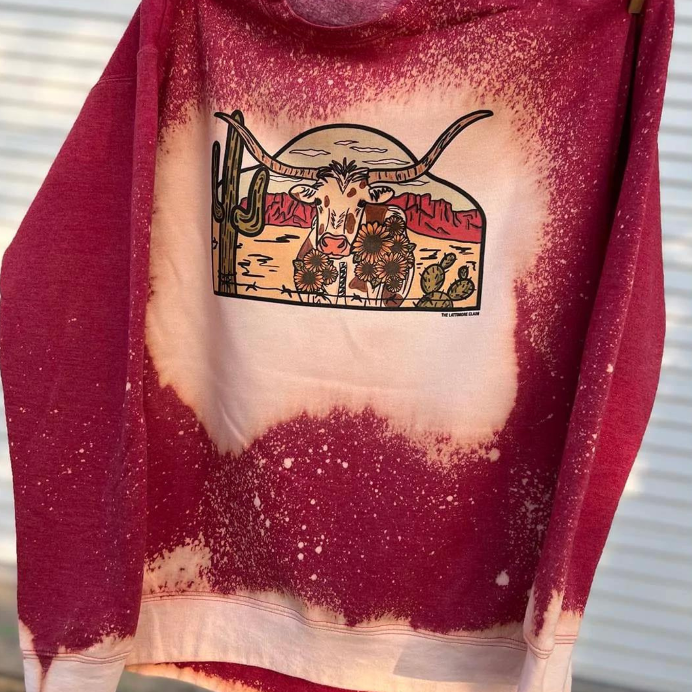 This bleached red graphic sweatshirt includes a crew neckline, long sleeves, and cute hand drawn design of a desert scene with a longhorn in the middle. This sweatshirt is shown being held up.  