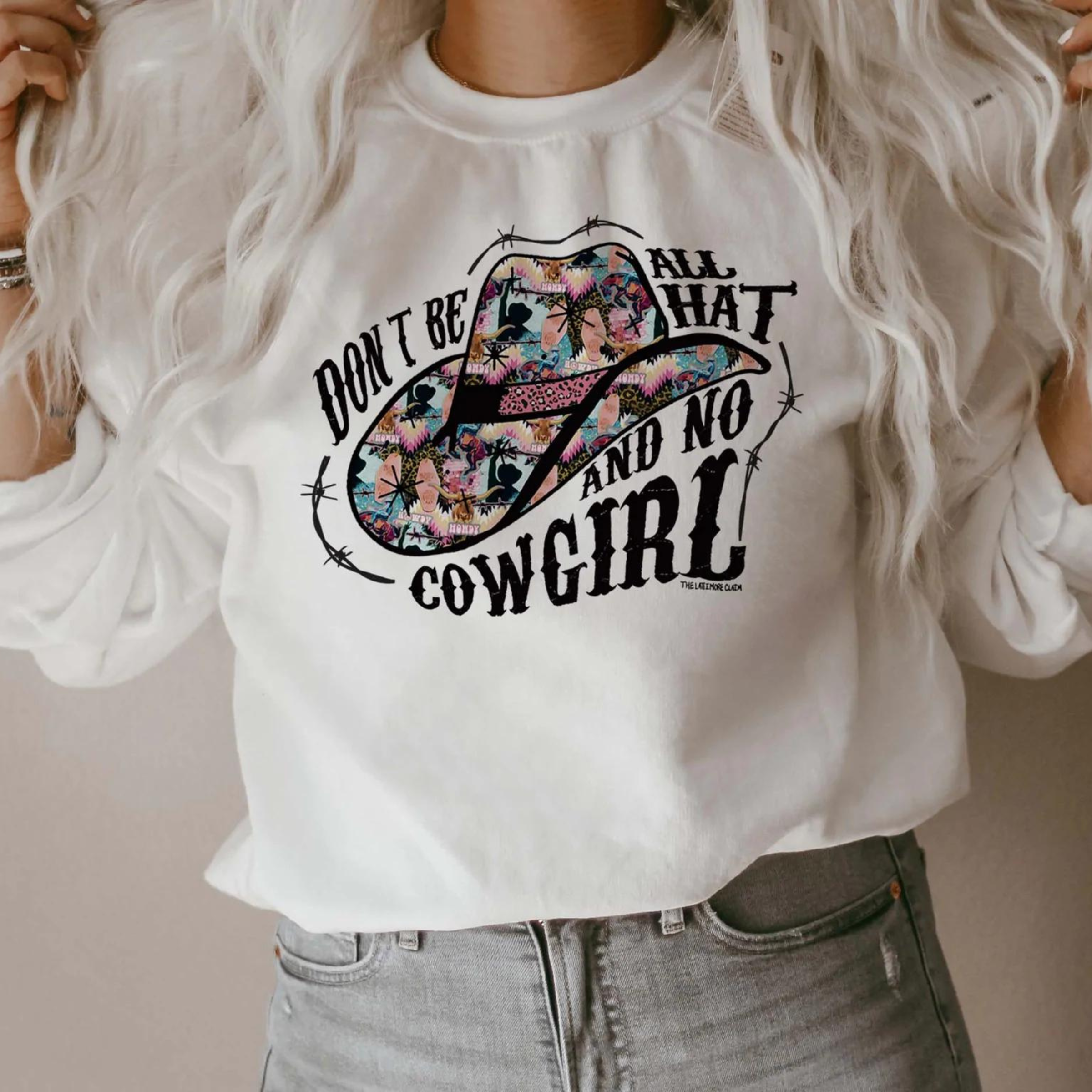 This white sweatshirt includes a crew neckline, long sleeves, and cute hand drawn design. of a cowboy hats when fun + bright prints, and the words "Don't Be All That And No Cowgirl". It also has black barbwire intertwined with the words and around the hat. This sweatshirt is shown in this photo to be styled with denim jeans and silver jewelry. The model has also rolled the sleeves. 