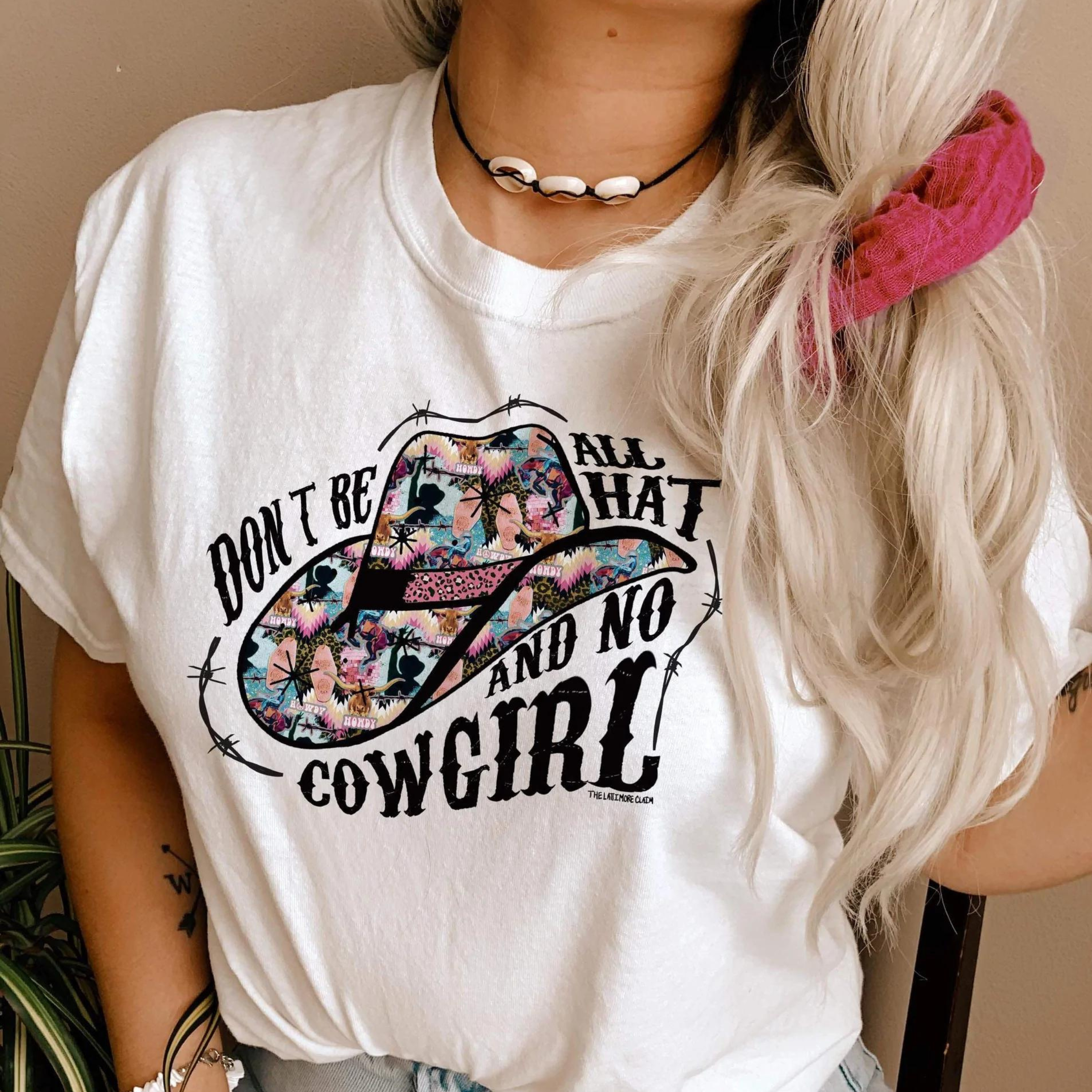 This white Bella + Canvas graphic tee includes a crew neckline, short sleeves, and cute hand drawn design. of a cowboy hats when fun + bright prints, and the words "Don't Be All That And No Cowgirl". It also has black barbwire intertwined with the words and around the hat. This sweatshirt is shown in this photo to be styled with denim jeans and shell jewelry. The model also has her hair pulled to the side with a pink scrunchie - fun pop of color! 