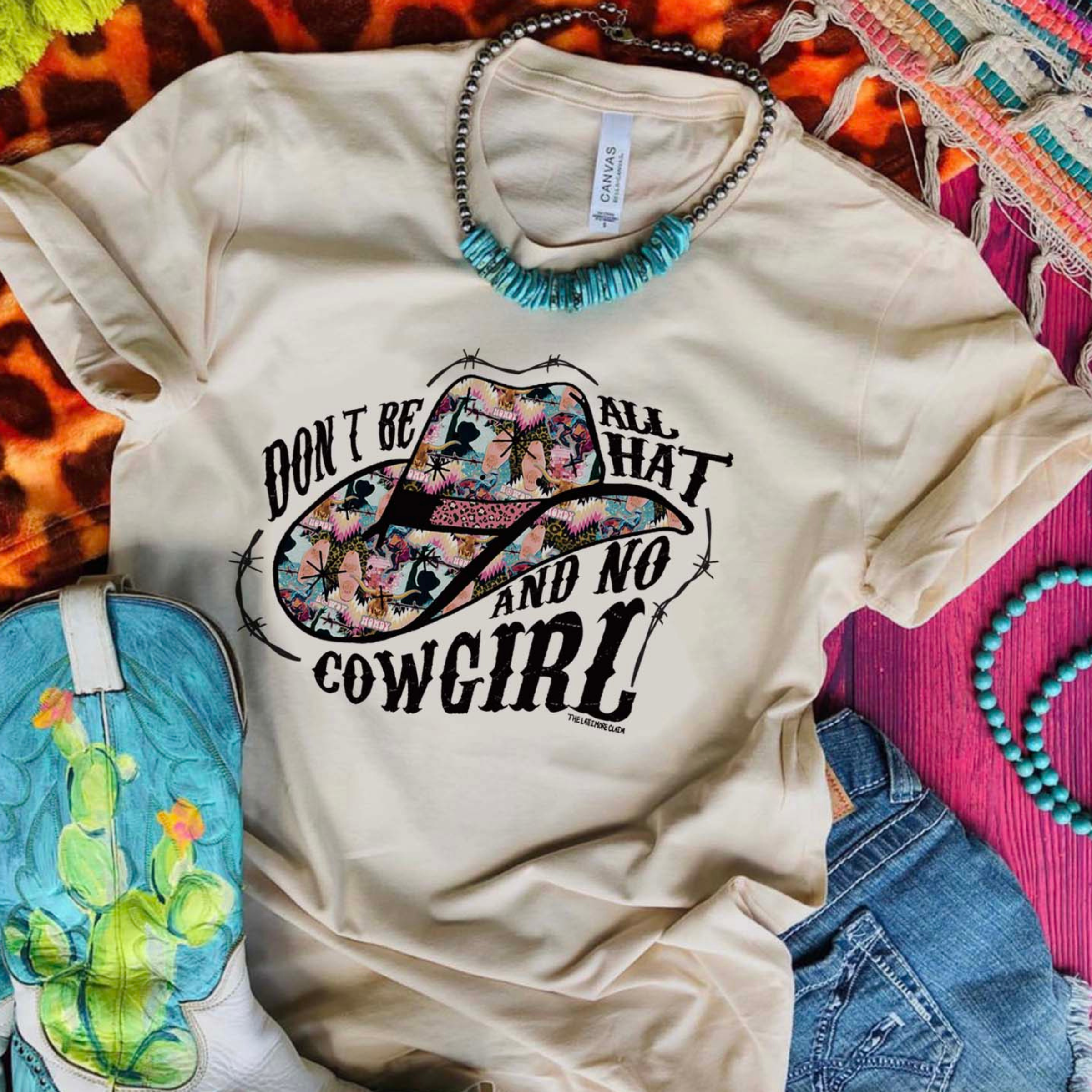 This cream Bella + Canvas graphic tee includes a crew neckline, short sleeves, and cute hand drawn design. of a cowboy hats when fun + bright prints, and the words "Don't Be All That And No Cowgirl". It also has black barbwire intertwined with the words and around the hat. This sweatshirt is shown in this photo to be styled as a flat lay with turquoise jewelry, denim jeans, and a pair of cowboy boots that have a cactus on them. 