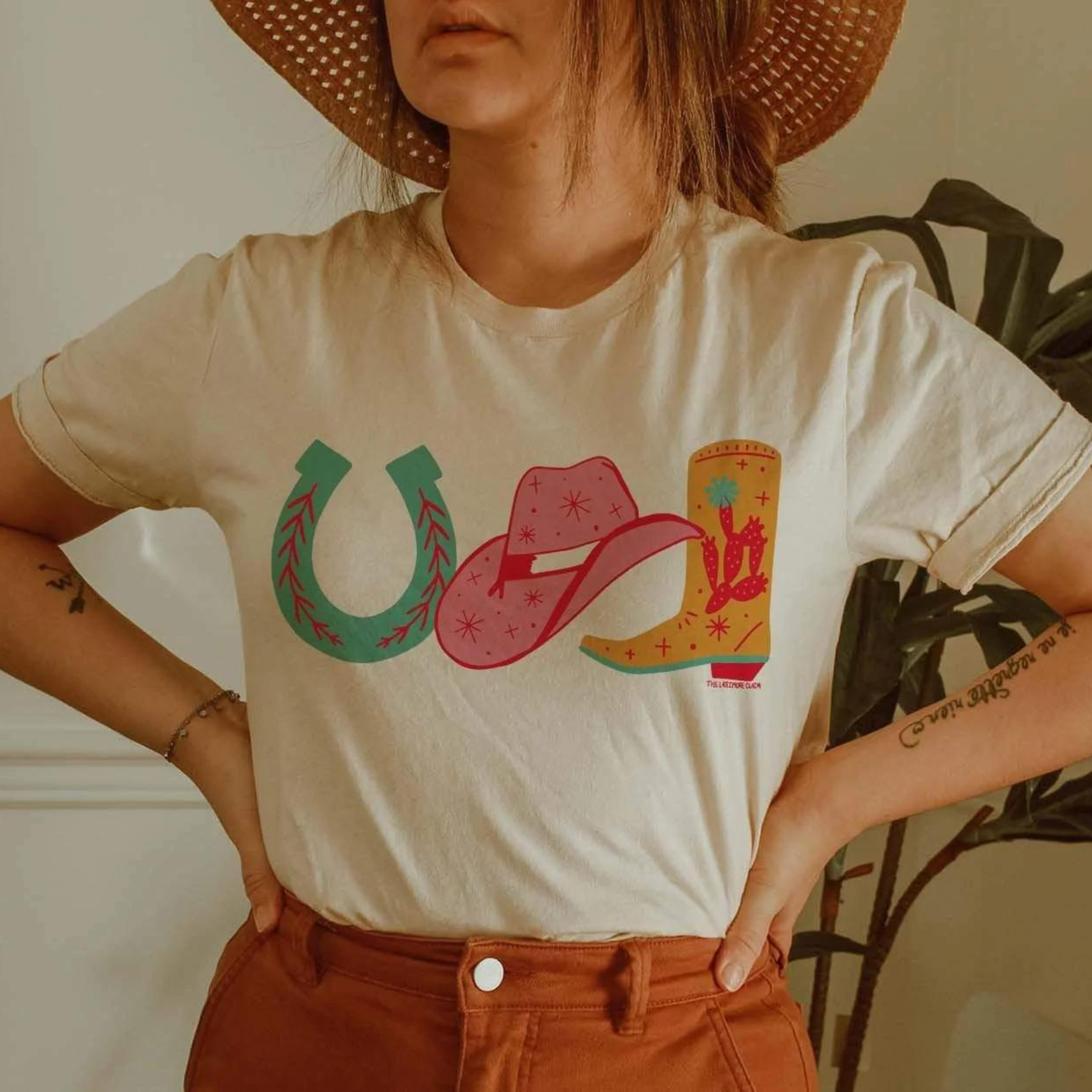 This cream graphic tee features three hand handdrawn cowgirl necessities - a horseshoe, a cowboy hat, and a boot in various pops of colors. This is a Bella + Canvas tee with short sleeves and a crew neckline. It is shown here styled with rust denim jeans, a silver bracelet, and a straw hat. 