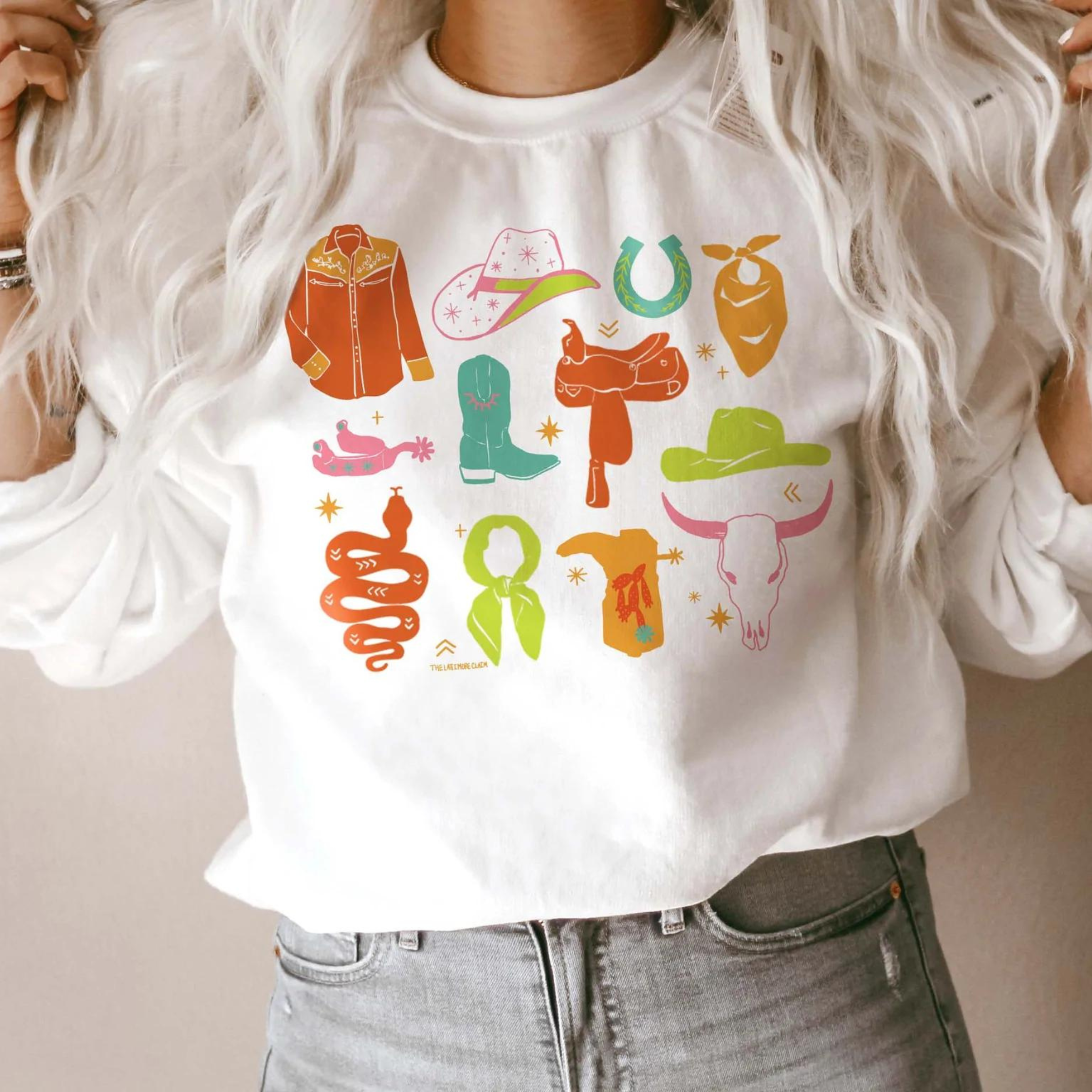 This white, hand-drawn graphic sweatshirt has a variety of cowgirl necessities and western finds. Items include a cowboy hat, a snake, a wild rag, and many more in various pops of colors. It is shown here styled with denim jeans and silver bracelets. This sweatshirt has long sleeves and a crew neckline. 