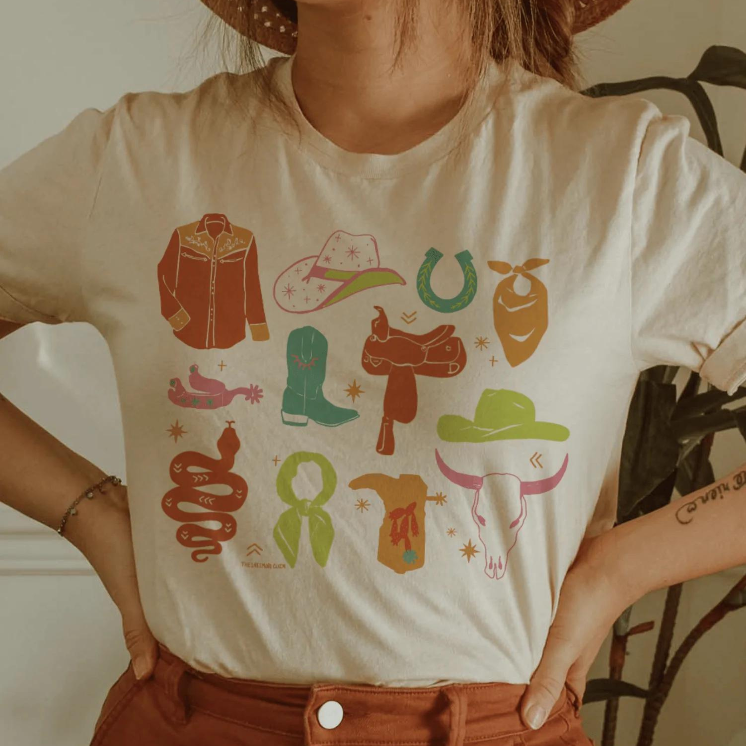 This cream, hand-drawn graphic tee has a variety of cowgirl necessities and western finds. Items include a cowboy hat, a snake, a wild rag, and many more in various pops of colors. This is a Bella + Canvas tee with short sleeves and a crew neckline. It is shown here styled with rust denim jeans, a straw hat, and a silver bracelet.
