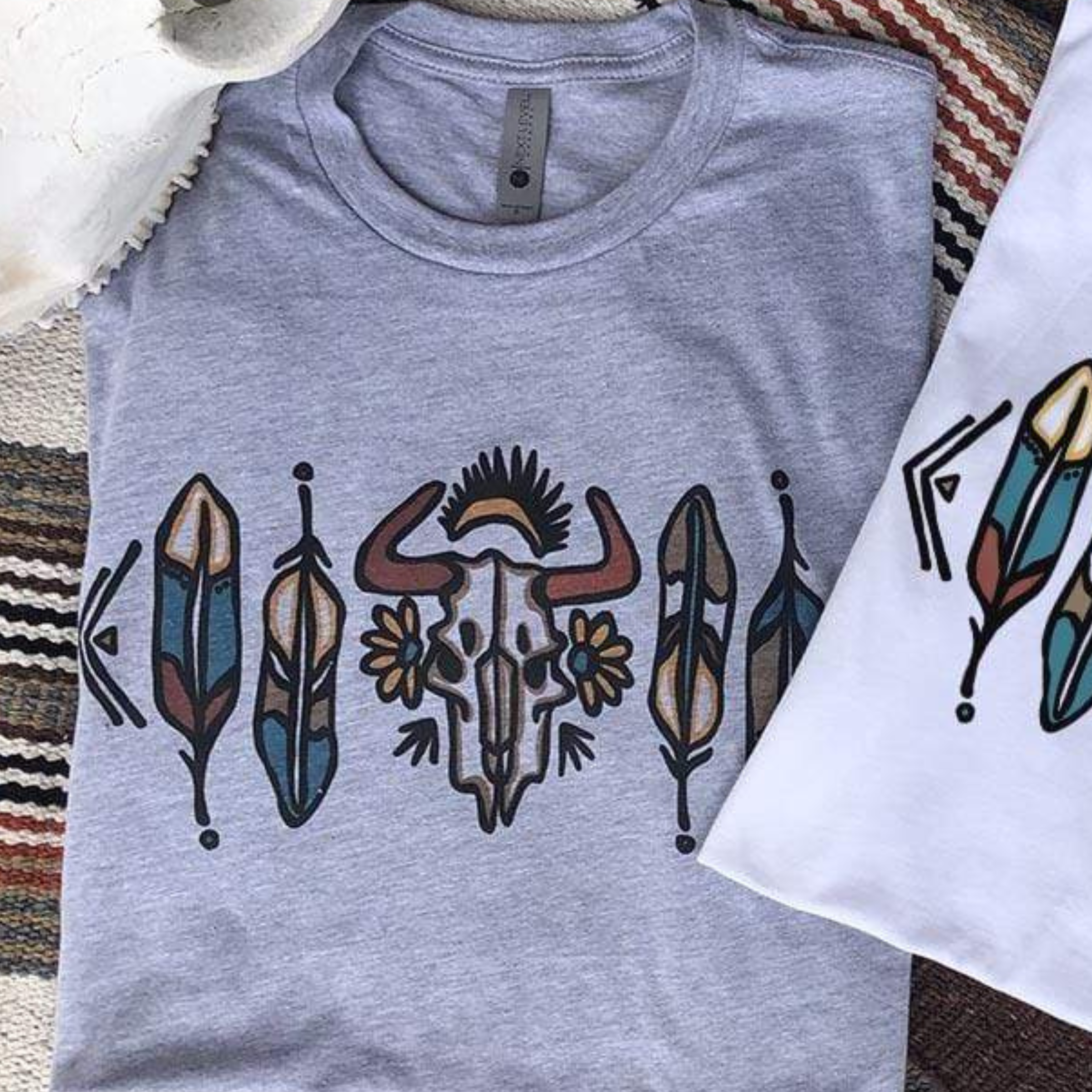 This Bella + Canvas tee in Grey includes a crew neckline, short sleeves, and a cute hand drawn design of a cow skull with two florals on either side along with 2 detailed feathers on both sides of the skull. The feathers include the following colors: blue, cream, dark green, and rust red.