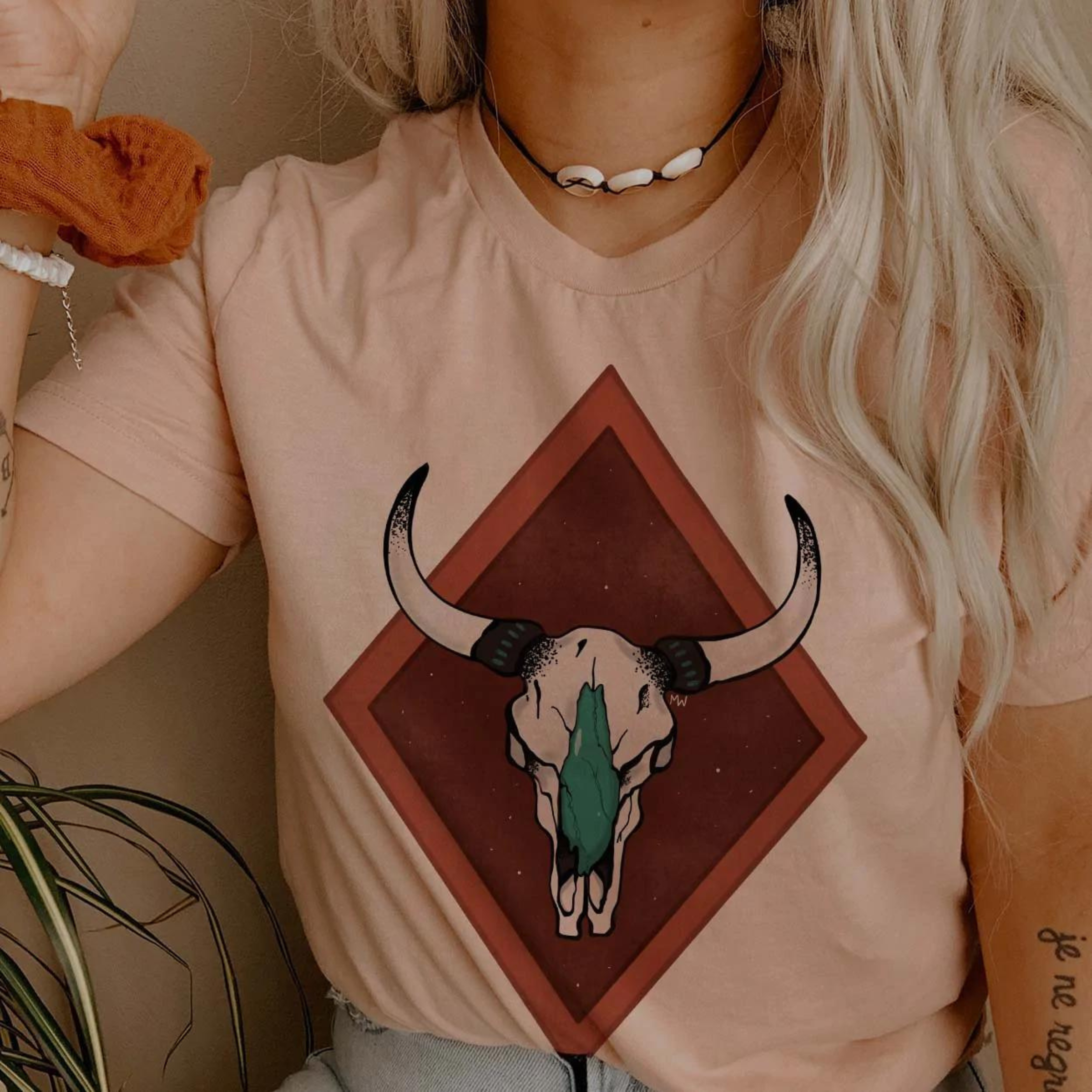 This Desert Rose Pink colored Bella + Canvas tee includes a crew neckline, short sleeves, and a cute hand drawn design of a detailed cow skull in white and turquoise, with a two-tone rust red double diamond behind it. 