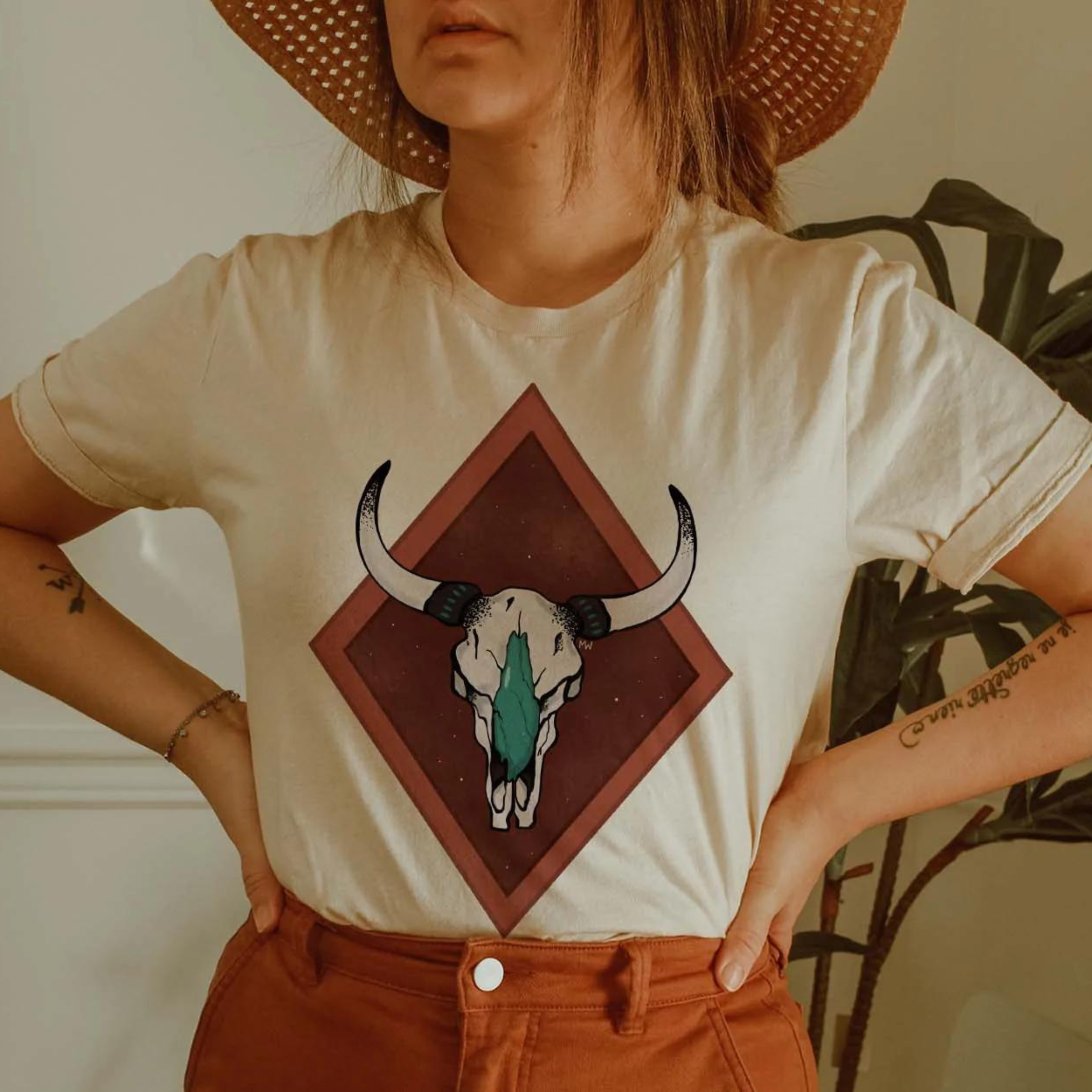 This Cream Bella + Canvas tee includes a crew neckline, short sleeves, and a cute hand drawn design of a detailed cow skull in white and turquoise, with a two-tone rust red double diamond behind it. 