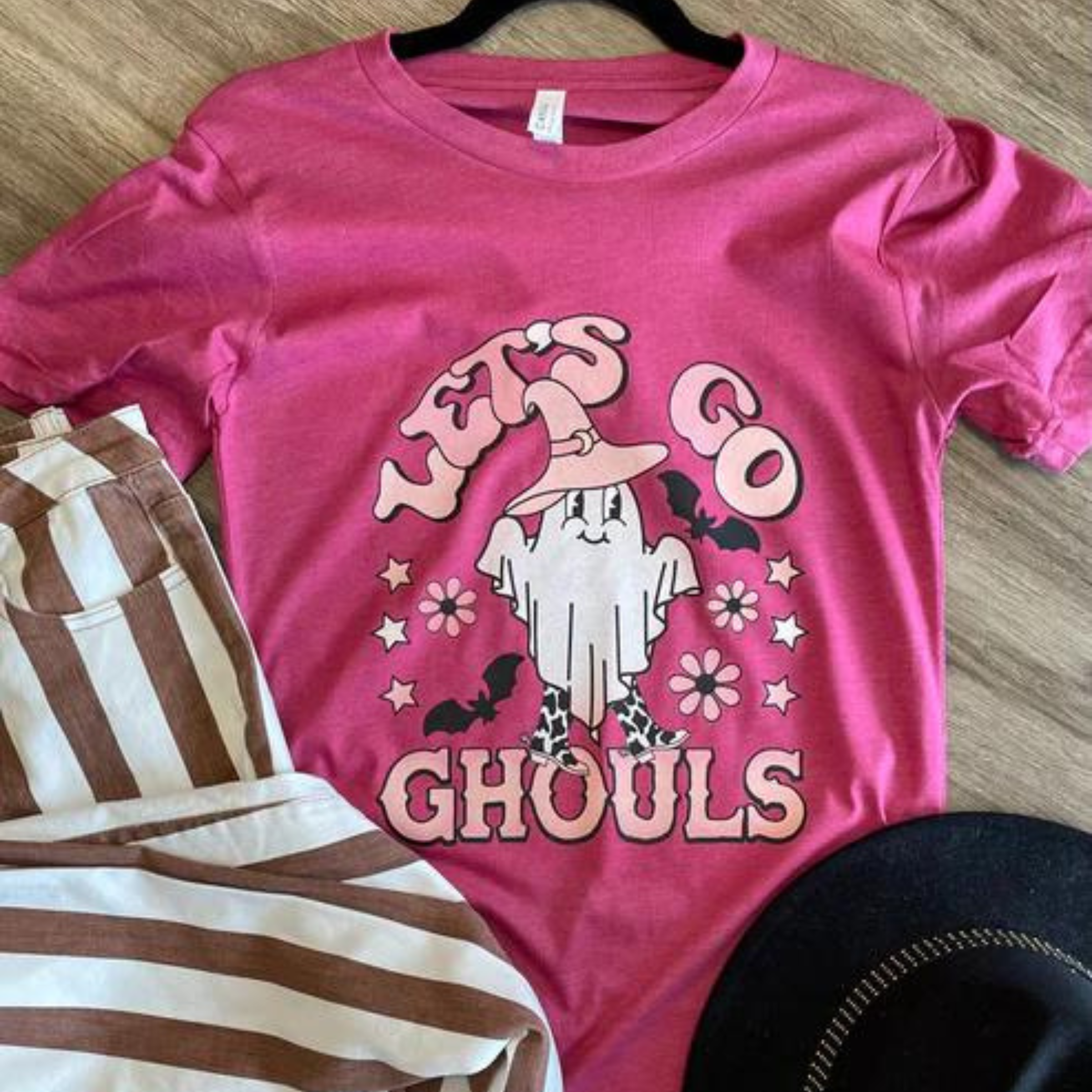 This pink Bella + Canvas tee includes a crew neckline, short sleeves, and a hand drawn graphic of a white ghost with a pink witch hat and cow print boots. The words "Let's Go Ghouls" are around the ghost along with two black bats, white and light pink stars, and three flowers in pink with black centers. "Let's Go Ghouls" is in a pretty, light pink. This is shown paired with a pair of brown and white vertical striped jeans and a black felt hat. 