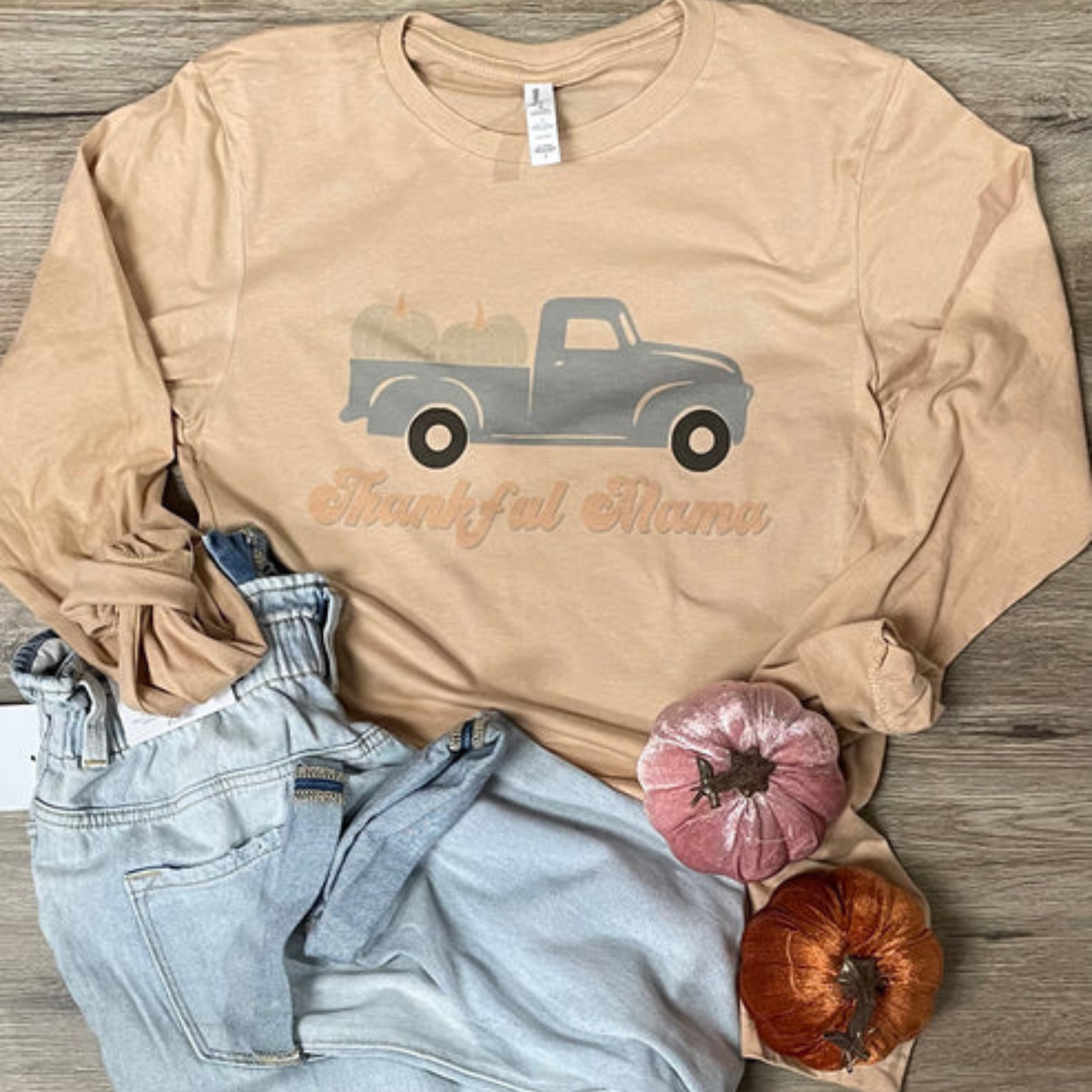 This cream long sleeve graphic tee from Bella + Canvas is crew neck and features a graphic of a Fall blue truck with two pumpkins in the end. Under the truck is the words "Thankful Mama" in pastel orange. This long sleeve graphic tee is shown paired with light wash denim jeans. 