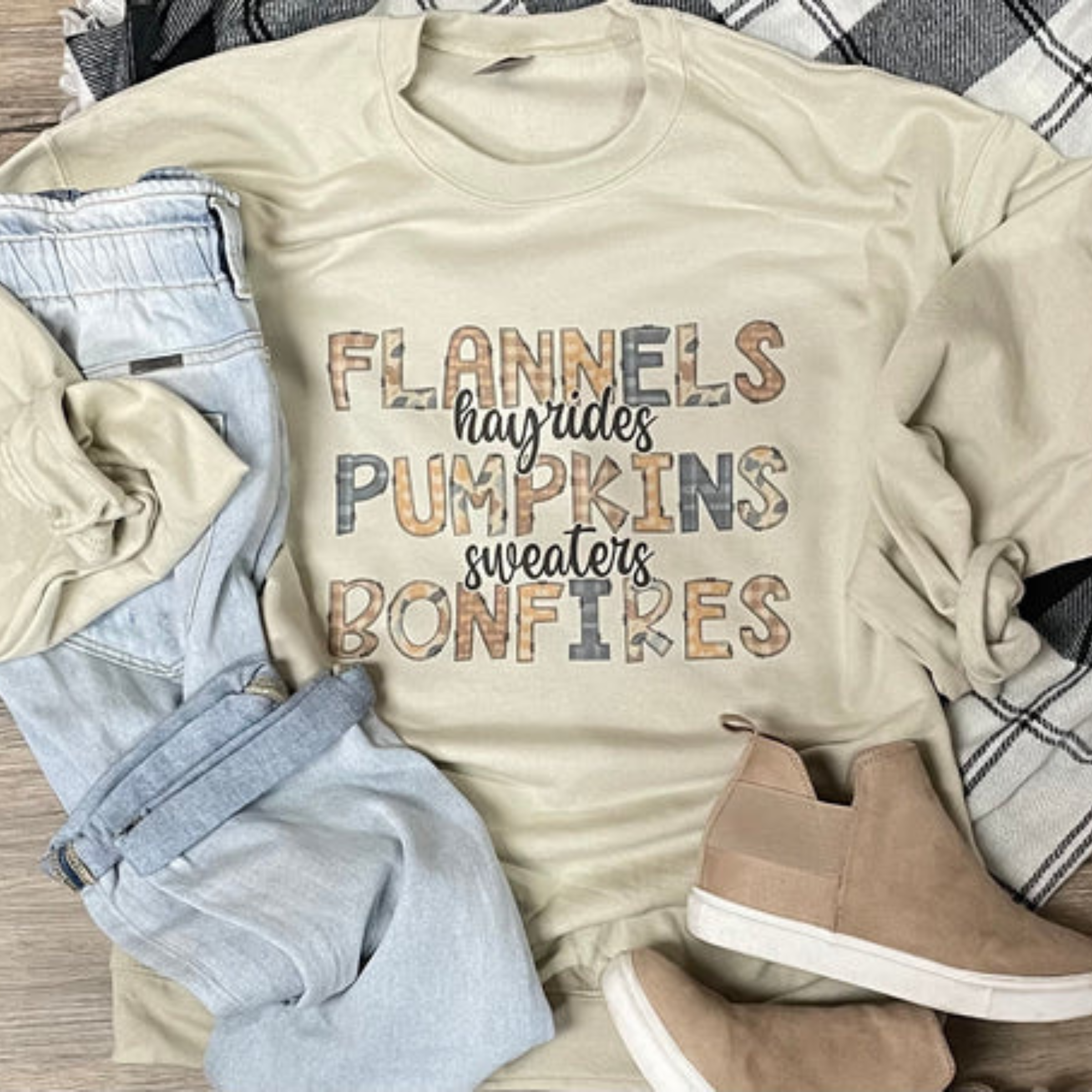 This cream sweatshirt features a hand drawn design that says "Flannels, Hayrides, Pumpkins, Sweaters, Bonfires". The words "Flannels", "Pumpkins", and "Bonfires" are in capital font with fun, Fall patterns. The words "Hayrides" and "Sweaters" are in a cursive, lowercase, black font. 