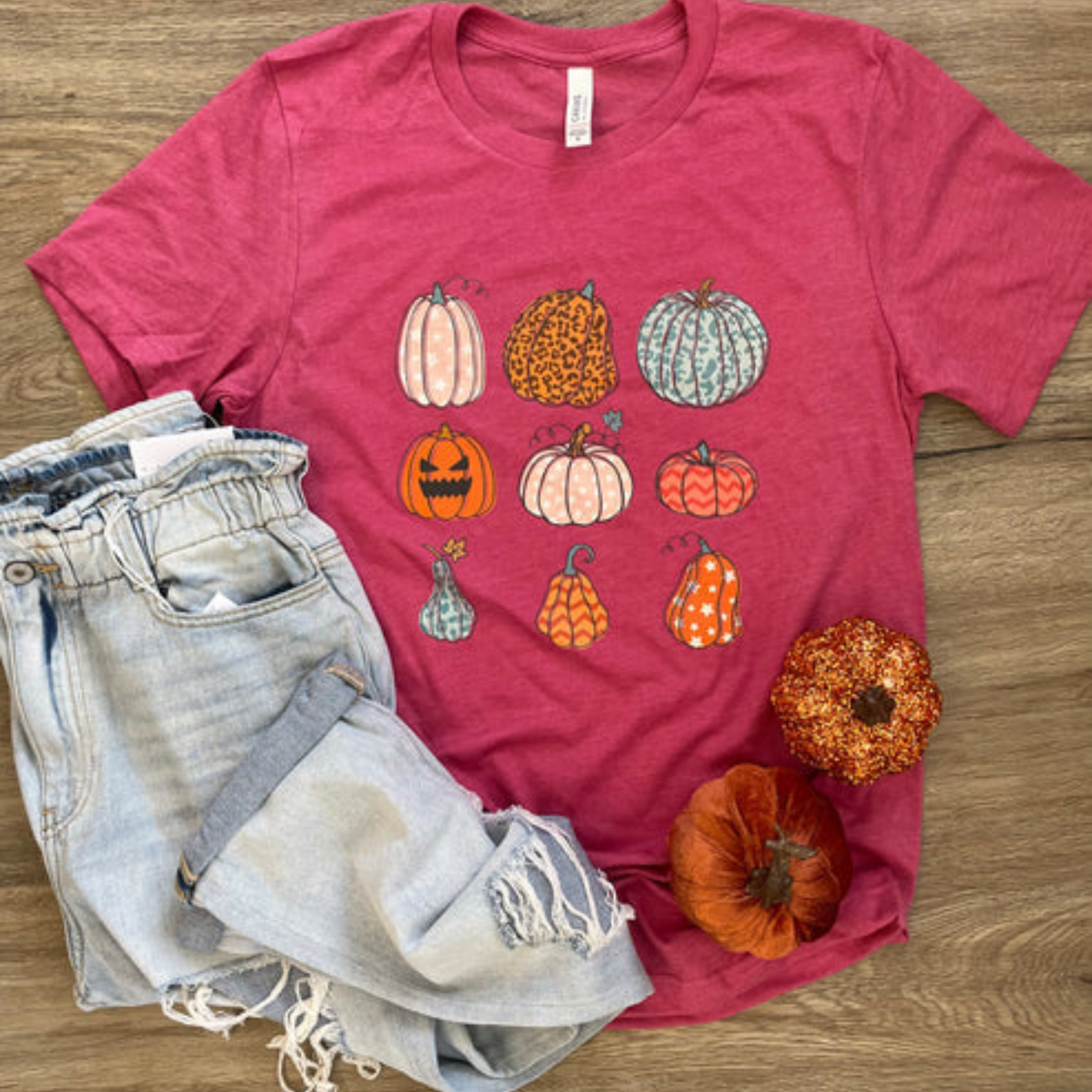 This heather red tee has a variety of pumpkins in different, seasonal pumpkins (nine pumpkins total, stacked in three rows of three). This is a Bella + Canvas tee and has short sleeves and a crew neckline. It is shown as a flat lay paired with light, distressed denim jeans. 