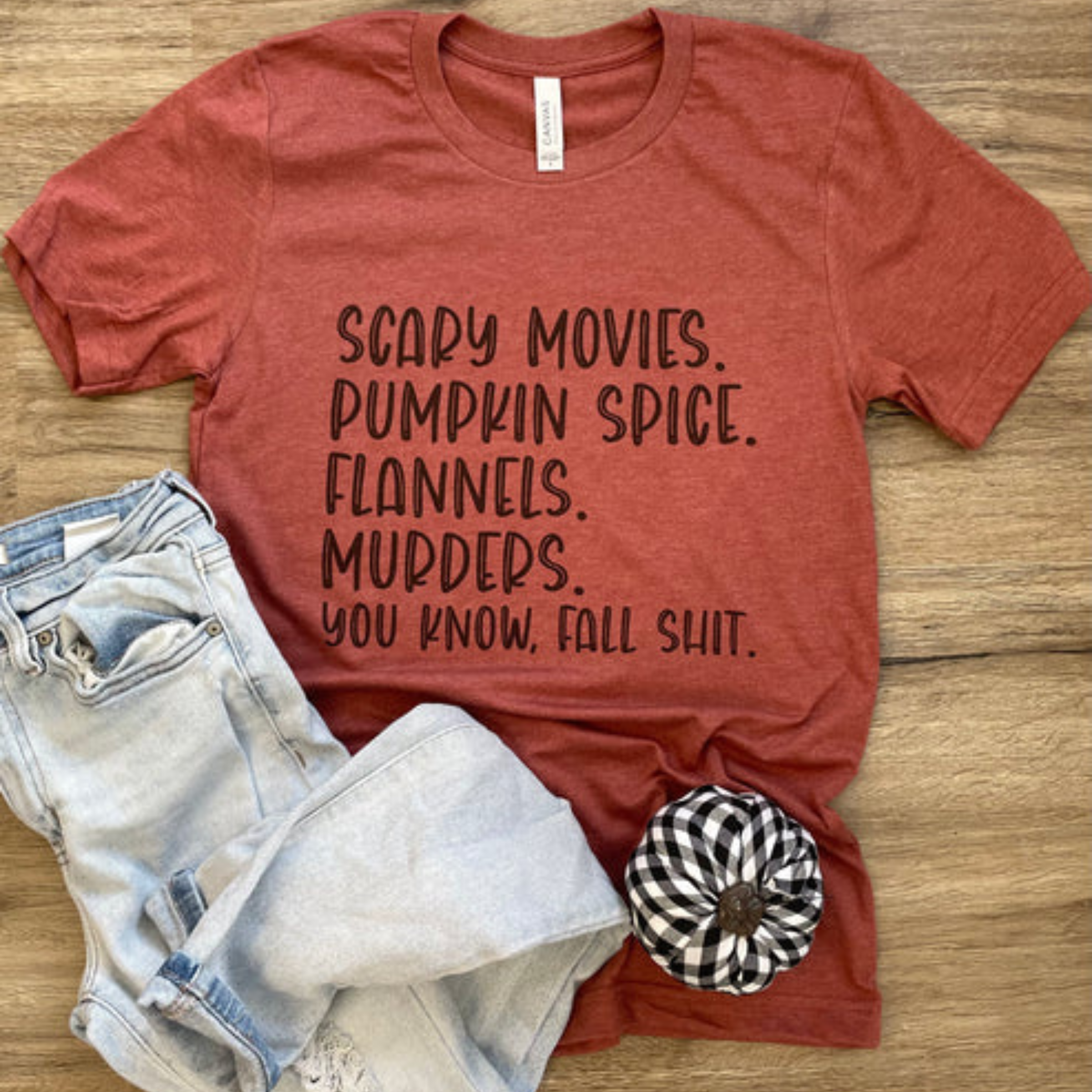 This heather rust red graphic tee says "Scary Movies. Pumpkin Spice. Flannels. Murders. You Know, Fall Shit." in a fun, black font stacked. This is a Bella + Canvas tee with short sleeves and a crew neckline. It is shown as a flat lay and paired with light, distressed denim jeans. 