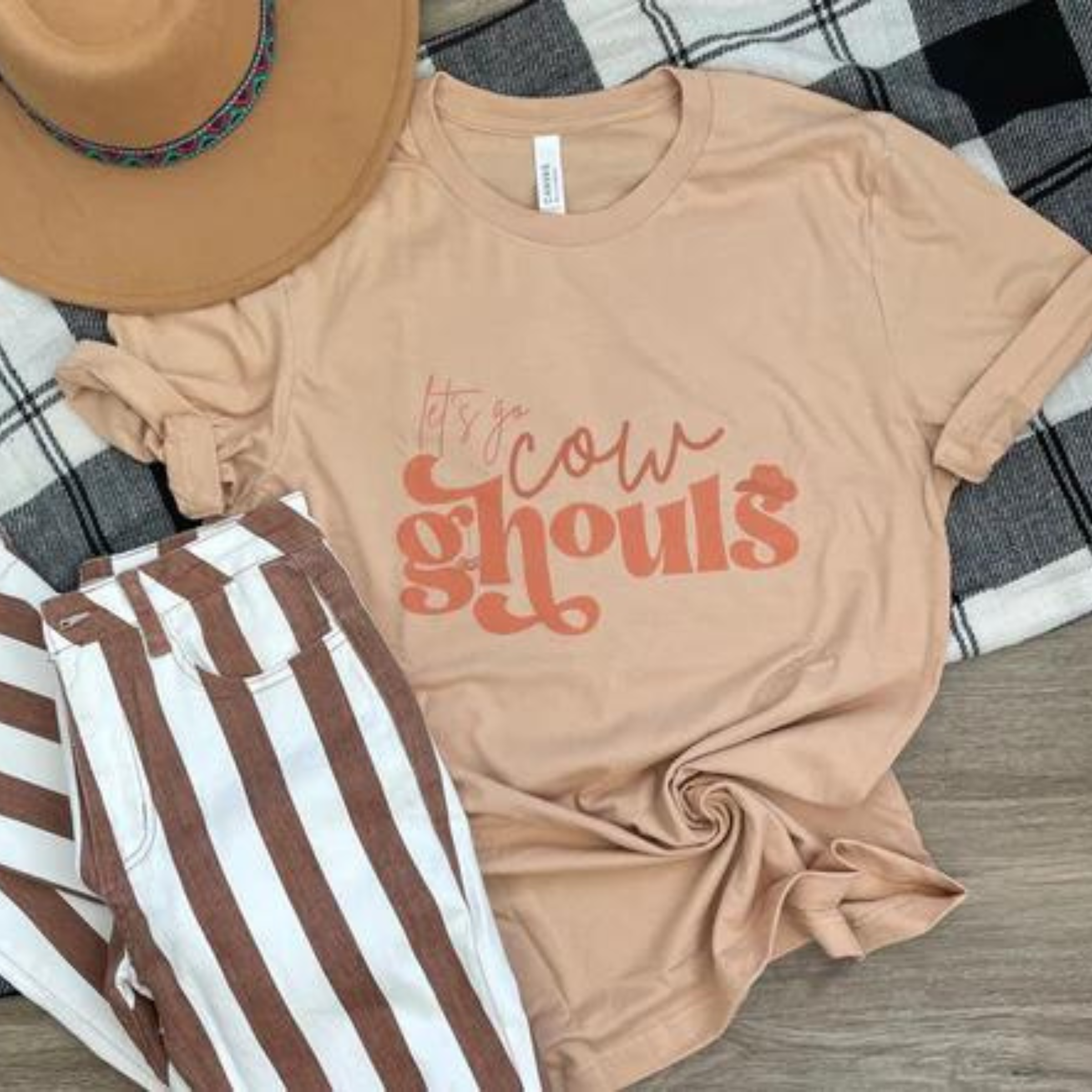 This tan graphic tee says "Let's Go Cow Ghouls" in orange, various fun fonts. This is a Bella + Canvas tee with short sleeves and crew neck style. It is shown as a flat lay and paired with a camel felt hat with a colorful hat band and brown and white vertical striped jeans. The sleeves of the tee are also shown folded up once. 