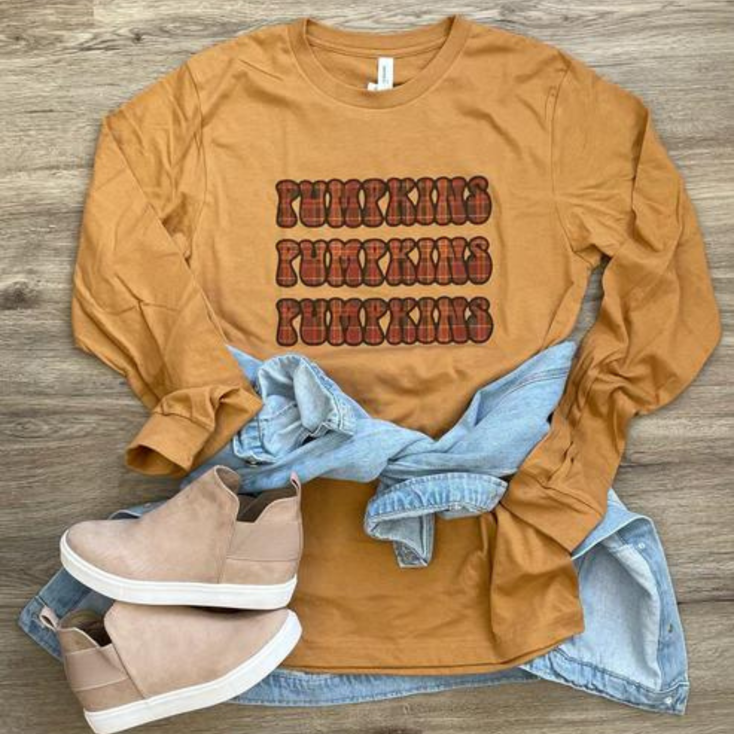 This mustard yellow graphic tee says "Pumpkins Pumpkins Pumpkins", which is in a flannel print and stacked. This is a Bella + Canvas long sleeve tee with a crew neck. It is shown as a flat lay with a light denim jacket tied around the waist and tan sneakers. 