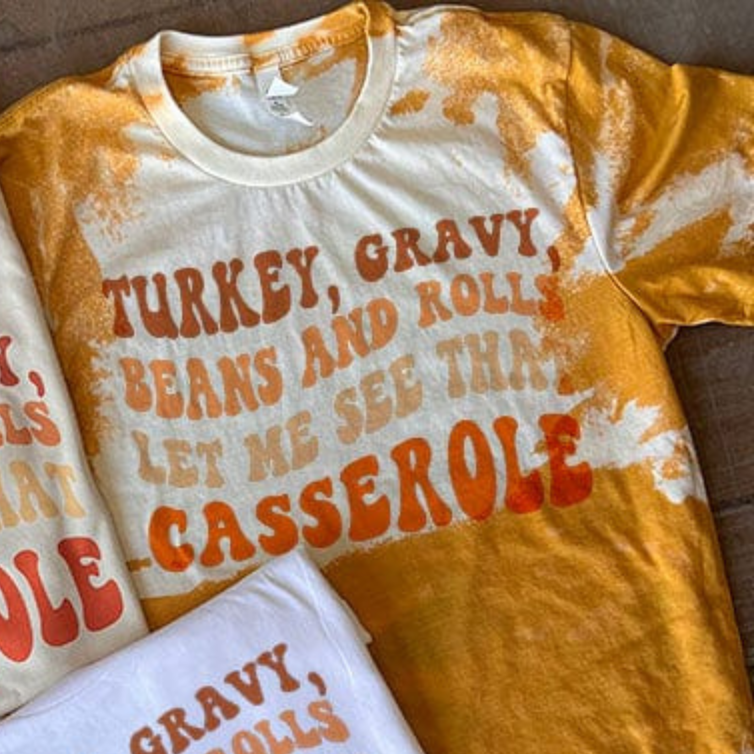 This bleached mustard yellow top includes a crew neckline, short sleeves, and a hand drawn graphic with words "Turkey, Gravy, Beans and Rolls, Let Me See That Casserole" in fun, Fall colors. This is a Bella + Canvas Tee, and shown here as a flat lay. 