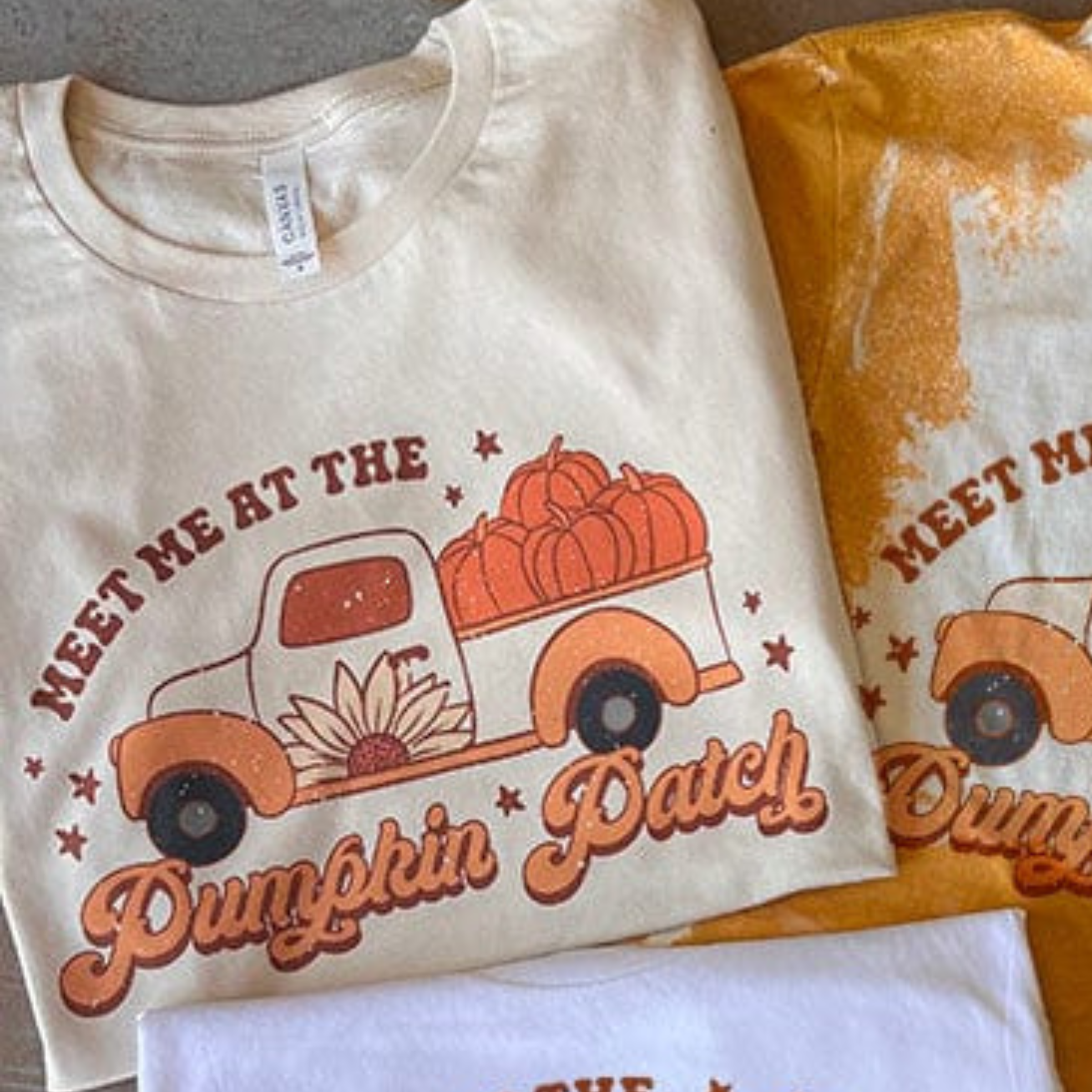 This cream top includes a crew neckline, short sleeves, and a hand drawn graphic with words "Meet Me At The Pumpkin Patch" around a Fall truck with pumpkins in the back. This is a Bella + Canvas Tee and styled in this photo as a folded flat lay.