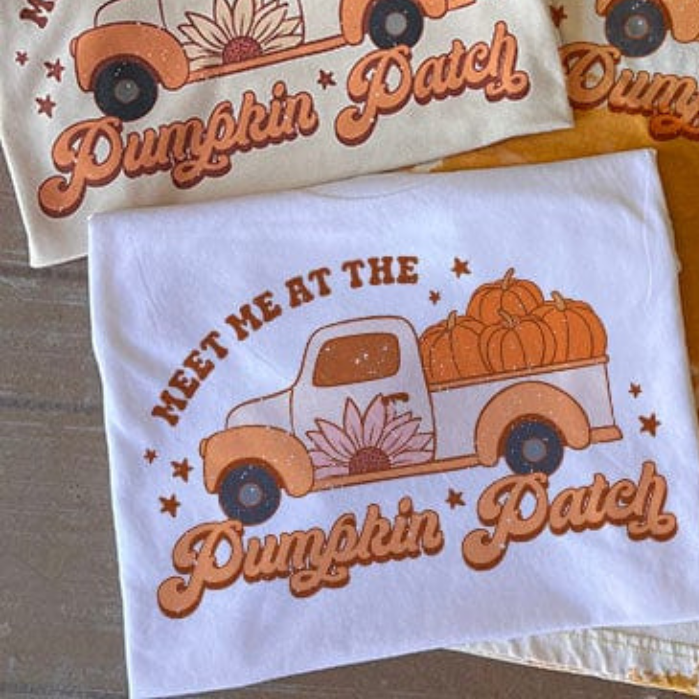 This white top includes a crew neckline, short sleeves, and a hand drawn graphic with words "Meet Me At The Pumpkin Patch" around a Fall truck with pumpkins in the back. This is a Bella + Canvas Tee and styled in this photo as a folded flat lay.