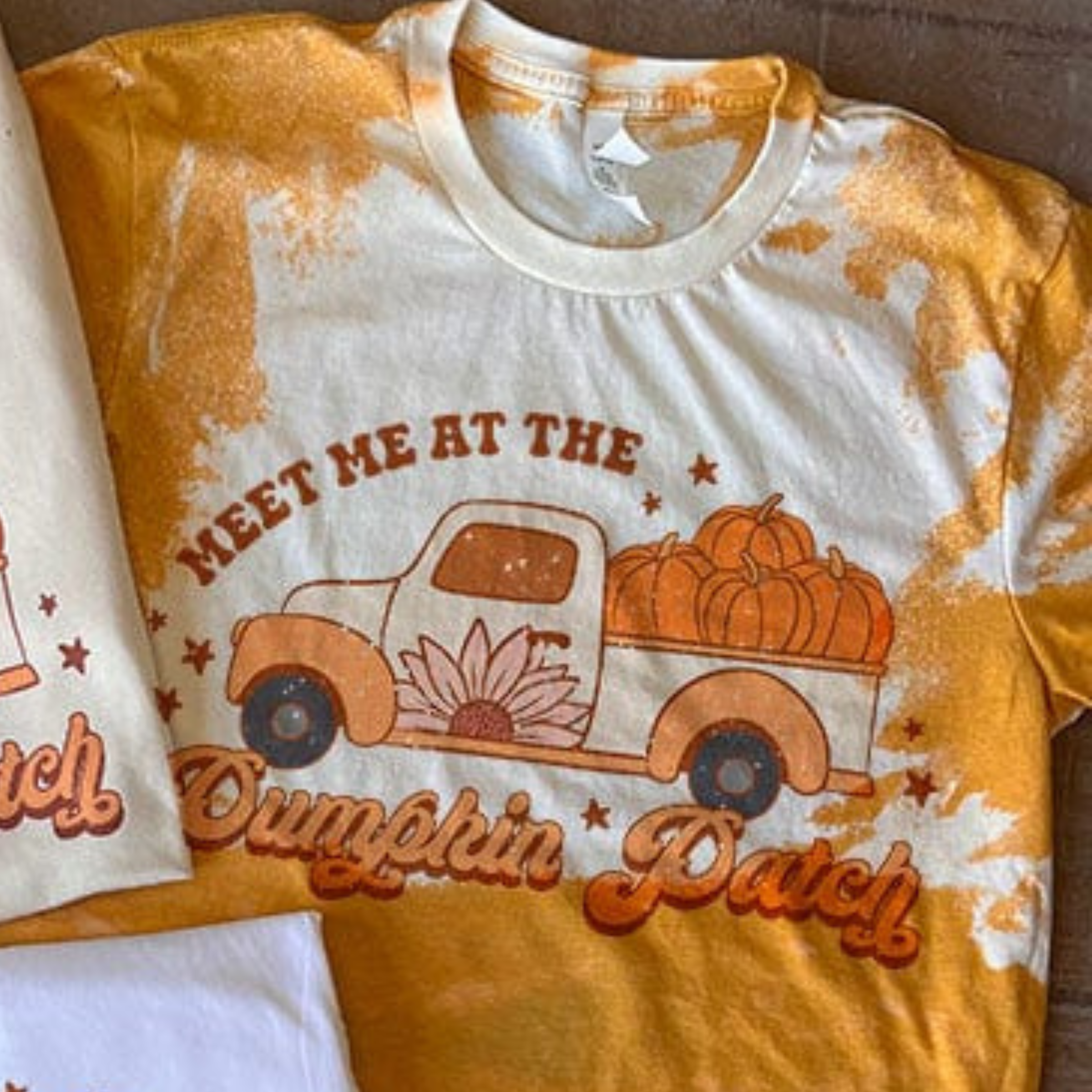 This bleached mustard yellow top includes a crew neckline, short sleeves, and a hand drawn graphic with words "Meet Me At The Pumpkin Patch" around a Fall truck with pumpkins in the back. This is a Bella + Canvas Tee and styled in this photo as a flat lay. 
