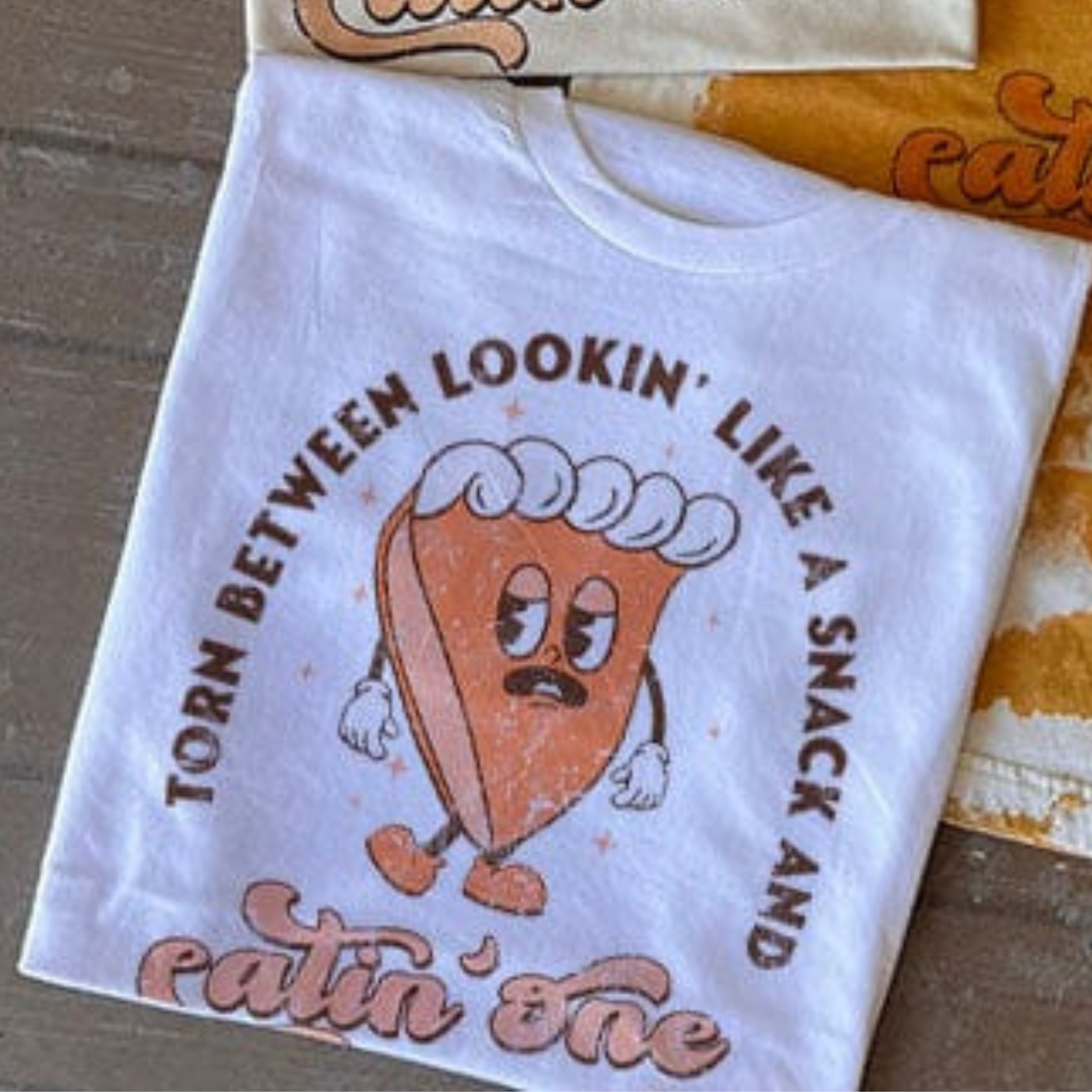 This white tee includes a crew neckline, short sleeves, and a cute hand drawn design of a slice of pie with the words "Torn Between Lookin' Like A Snack And Eatin' One" around it. It is shown styled as a folded flat lay.