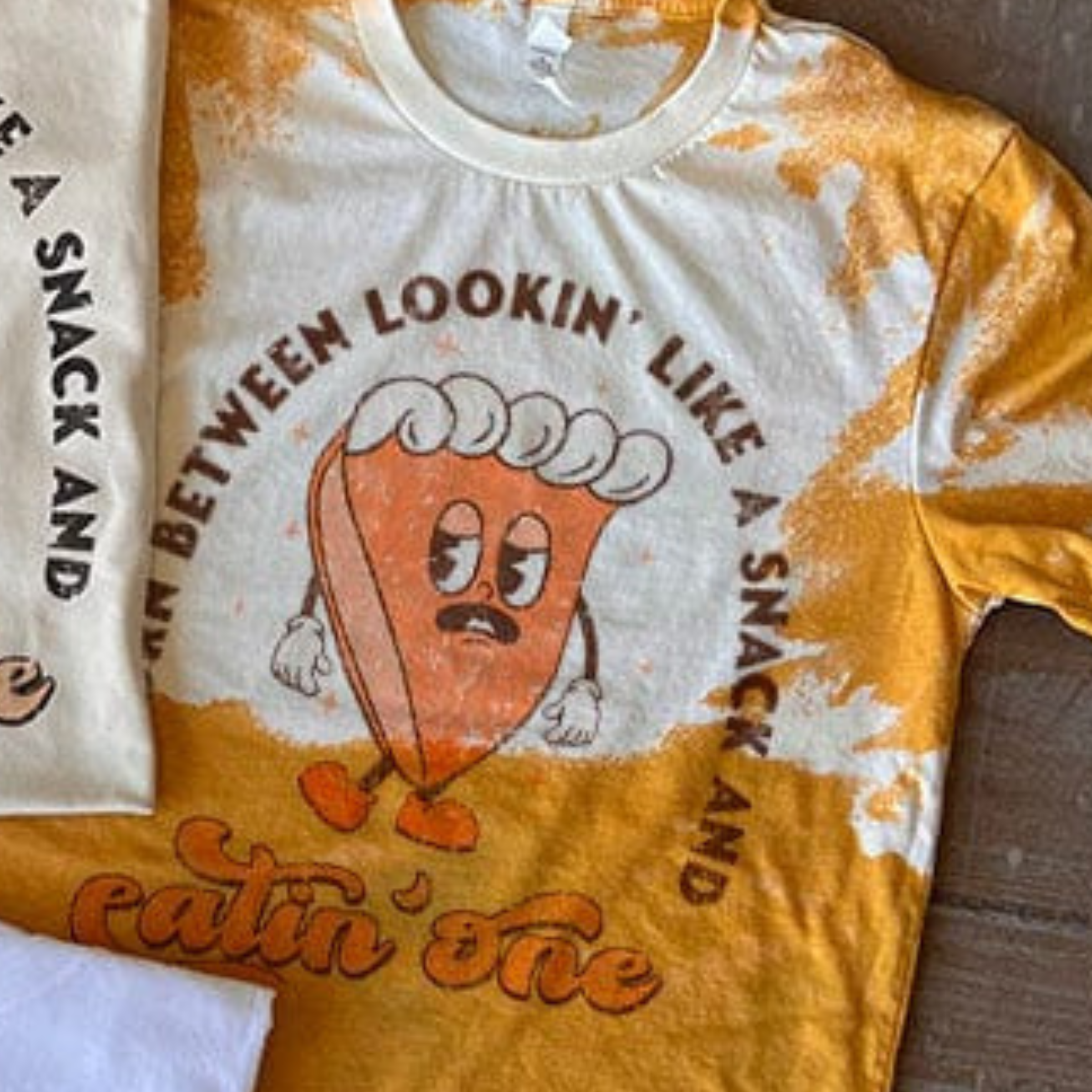 This bleached mustard yellow tee includes a crew neckline, short sleeves, and a cute hand drawn design of a slice of pie with the words "Torn Between Lookin' Like A Snack And Eatin' One" around it. It is shown styled as a flat lay in this photo.