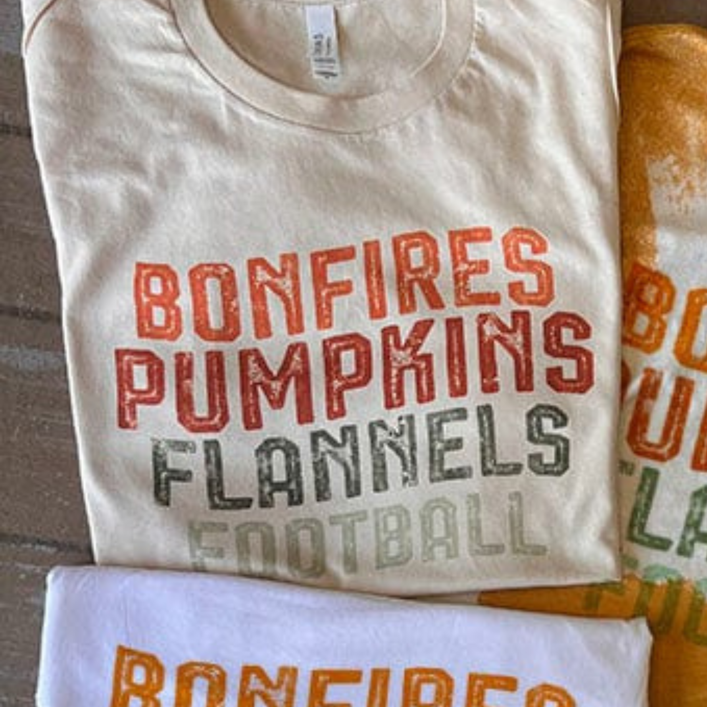 This cream tee includes a crew neckline, short sleeves, and a graphic with words "Bonfires Pumpkins Flannels Football". This is a Bella + Canvas Tee. It is shown as a folded flat lay in this photo.  