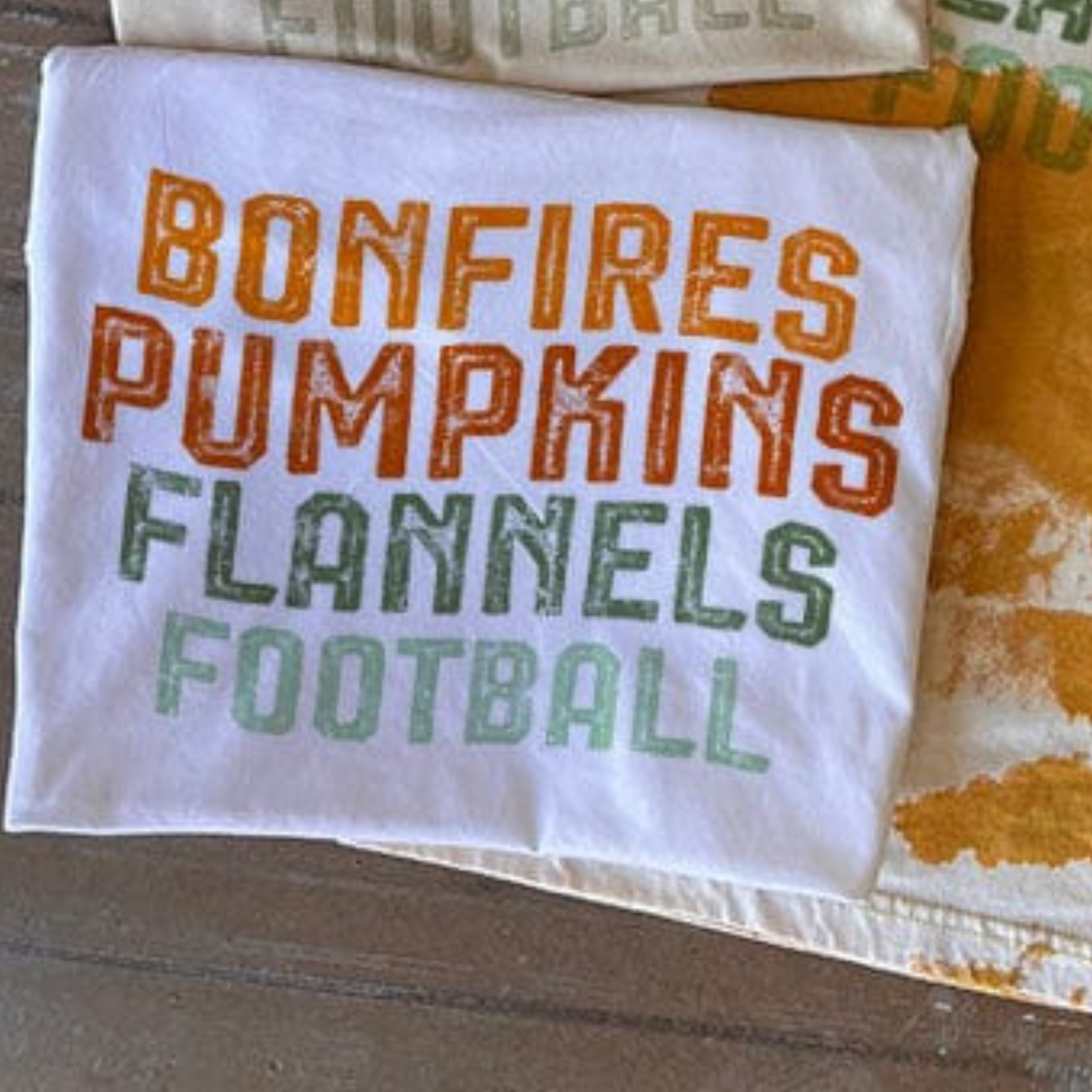 This white tee includes a crew neckline, short sleeves, and a graphic with words "Bonfires Pumpkins Flannels Football". This is a Bella + Canvas Tee. It is shown in this photo as a folded flat lay. 