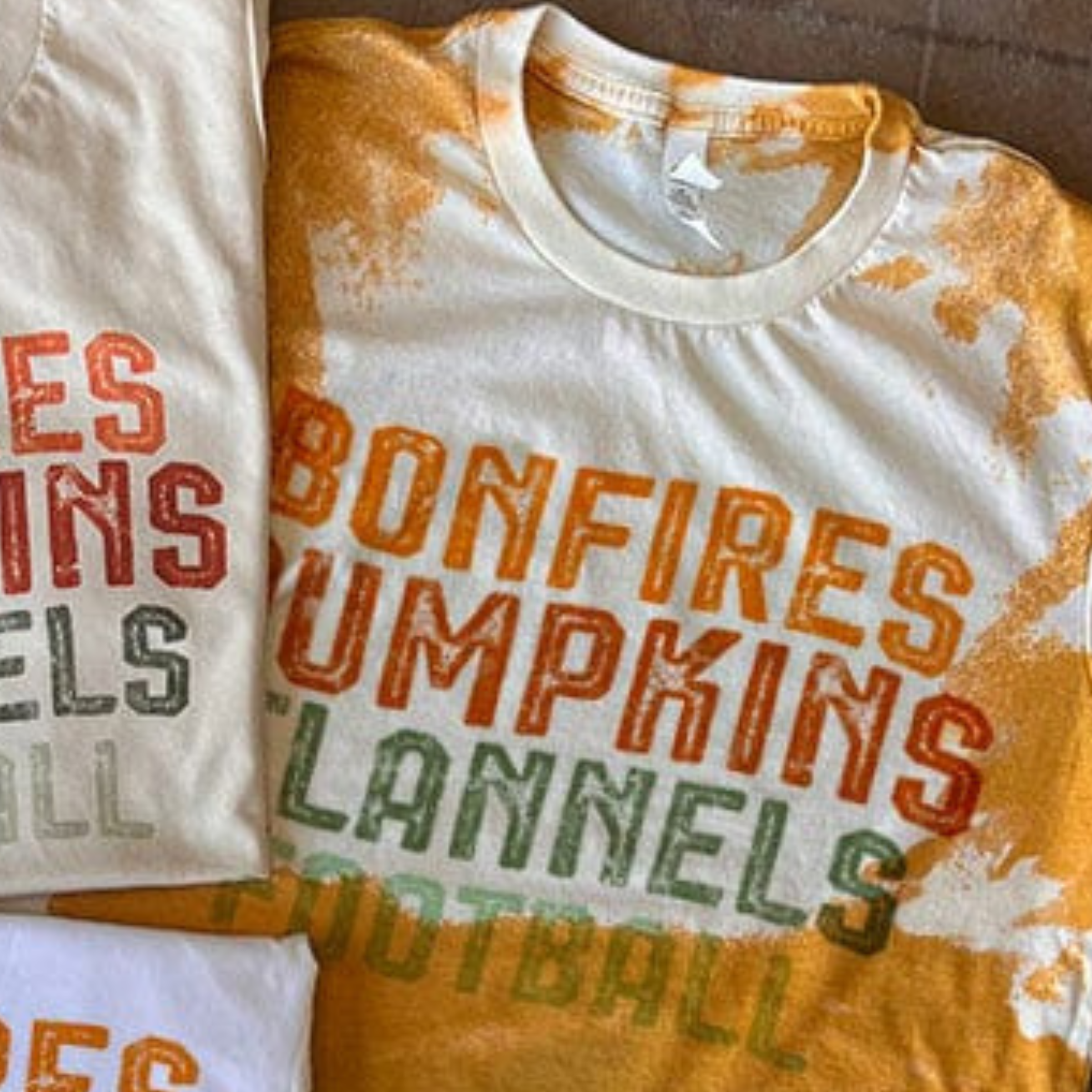 This bleached mustard yellow tee includes a crew neckline, short sleeves, and a graphic with words "Bonfires Pumpkins Flannels Football". This is a Bella + Canvas Tee. It is shown as a flat lay in this photo. 