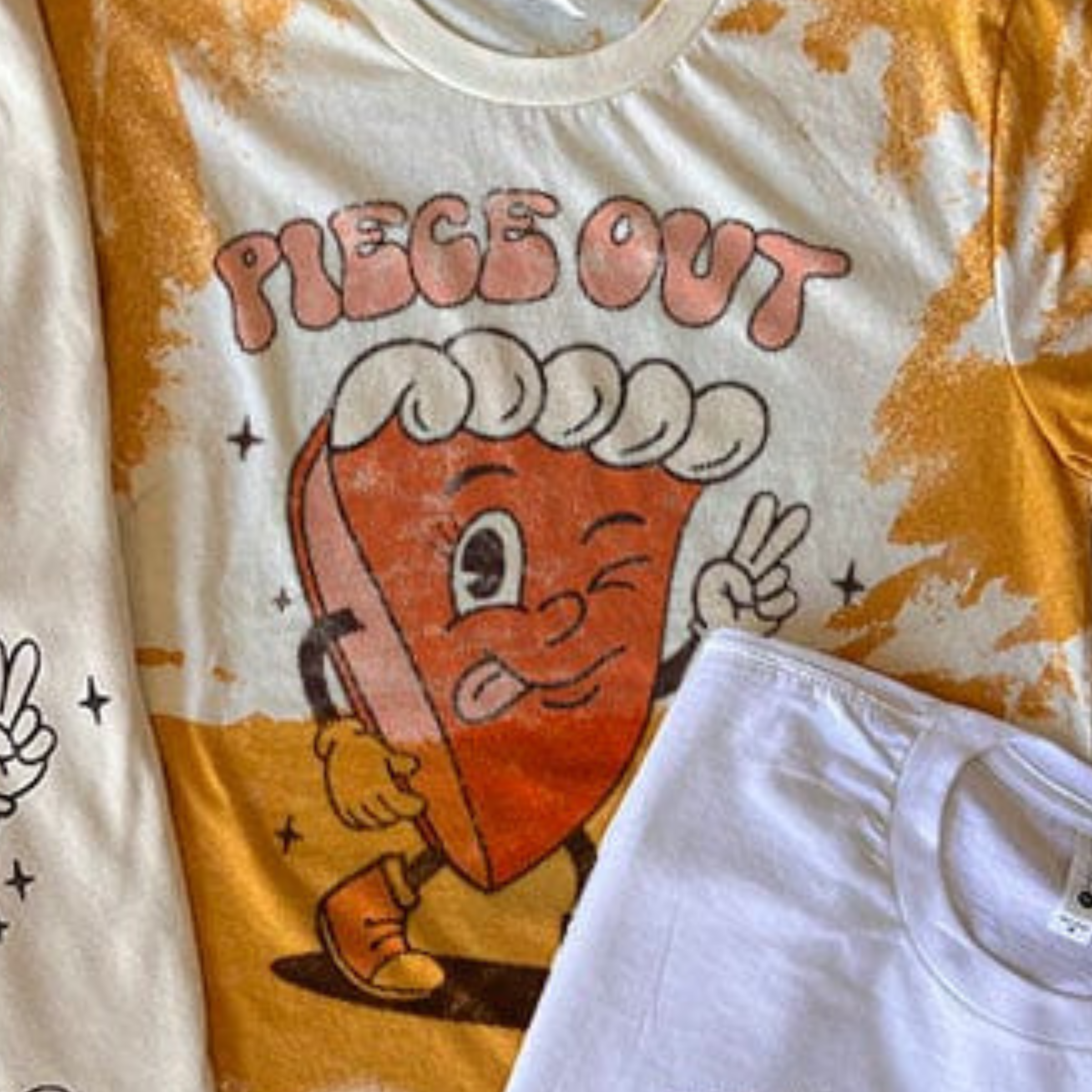 This bleached mustard yellow graphic tee includes a crew neckline, short sleeves, and a cute hand drawn design of a piece of pie with hands, legs, and feet. The pie's left hand is holding up the peace sign with the words "Piece Out" above it. This is shown as a flat lay in this photo. 