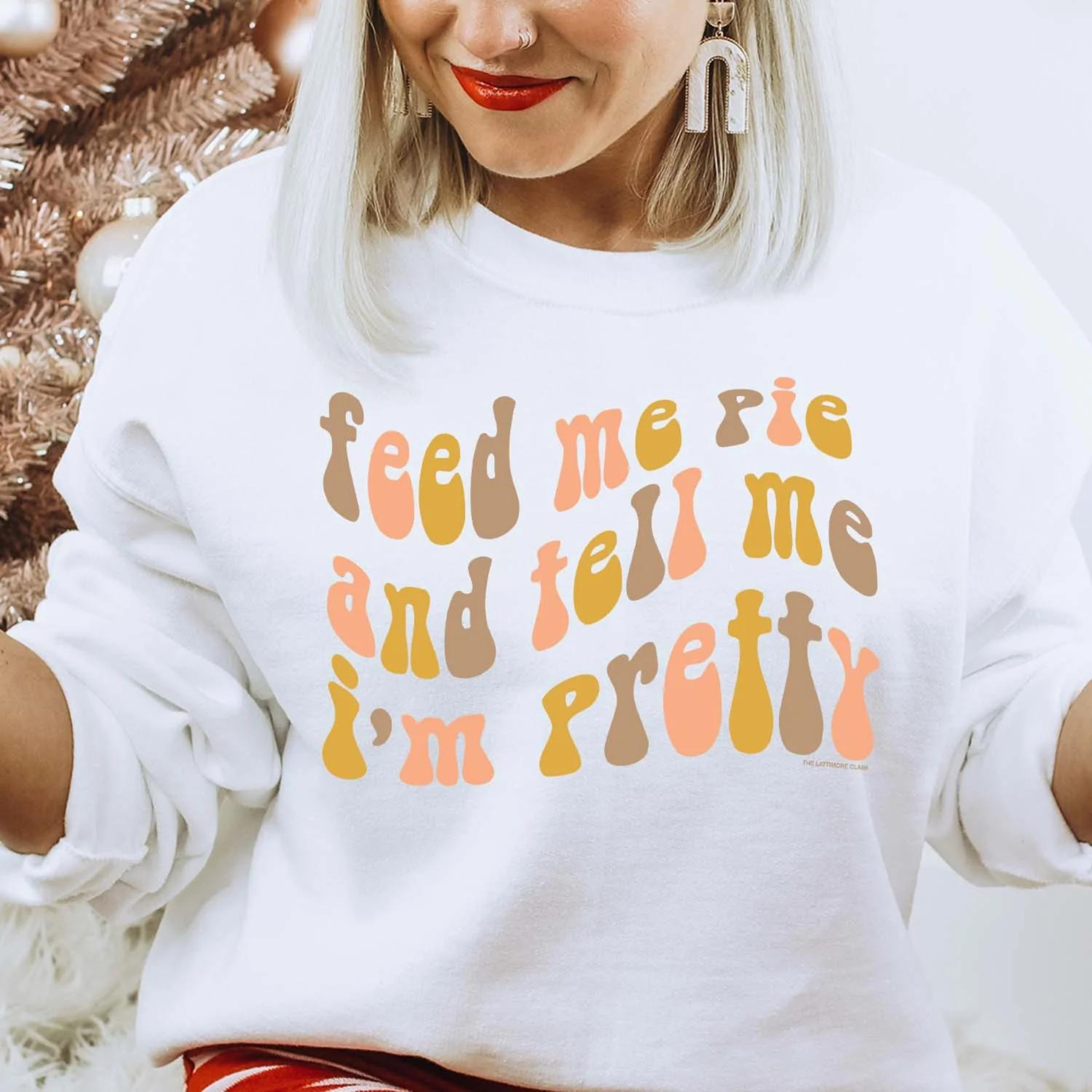 Model is wearing a white sweatshirt saying "feed me pie and tell me I'm pretty" with colors alternating in brown, pink, and mustard. Model has white dangle earrings. Background is white with a brown Christmas tree behind the model.