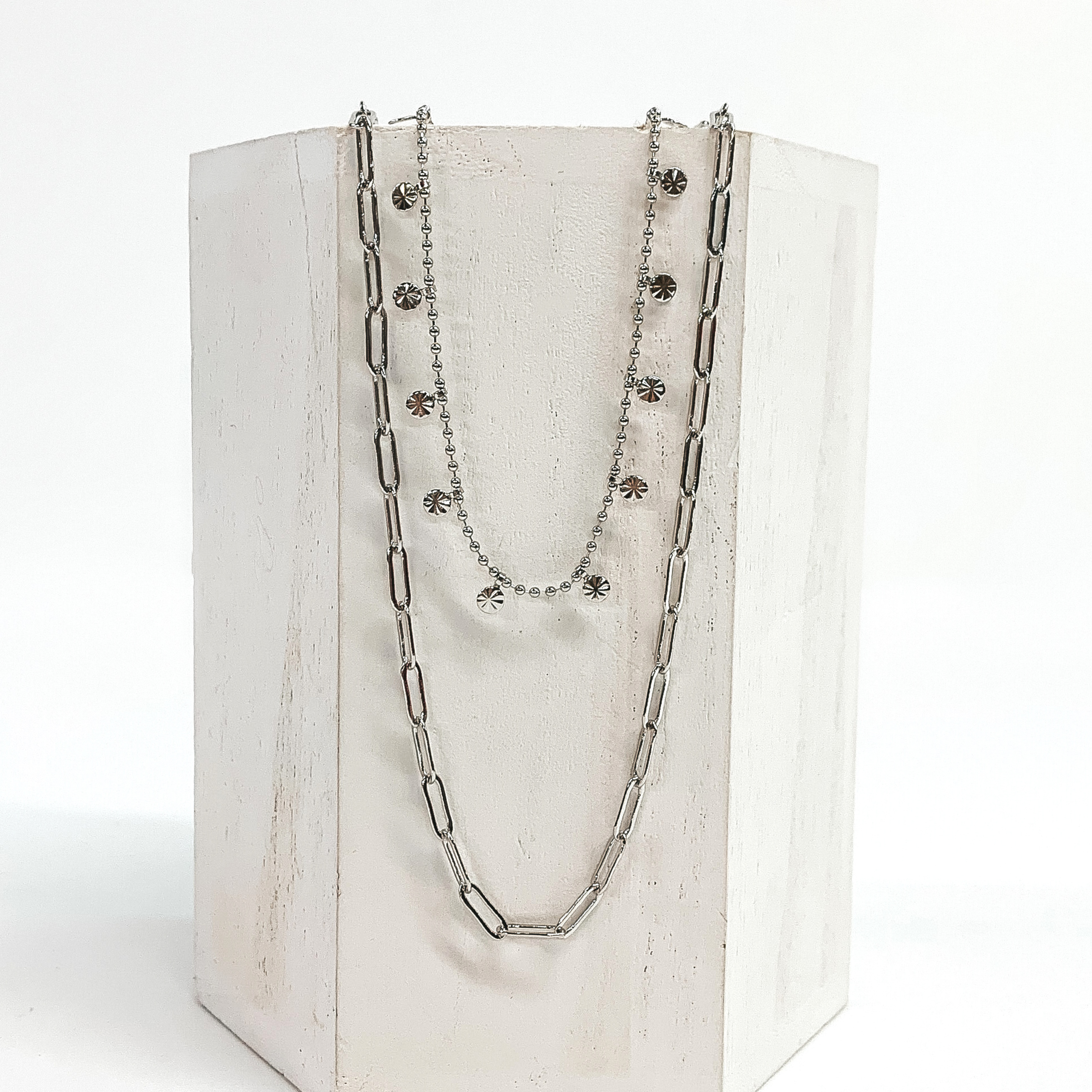 This is a silver double layered necklace. One strand is a thin paperclip chain and the second chain is a beaded chain with tiny charms with a sunburst design. This necklace is pictured on a white block and on a white background.