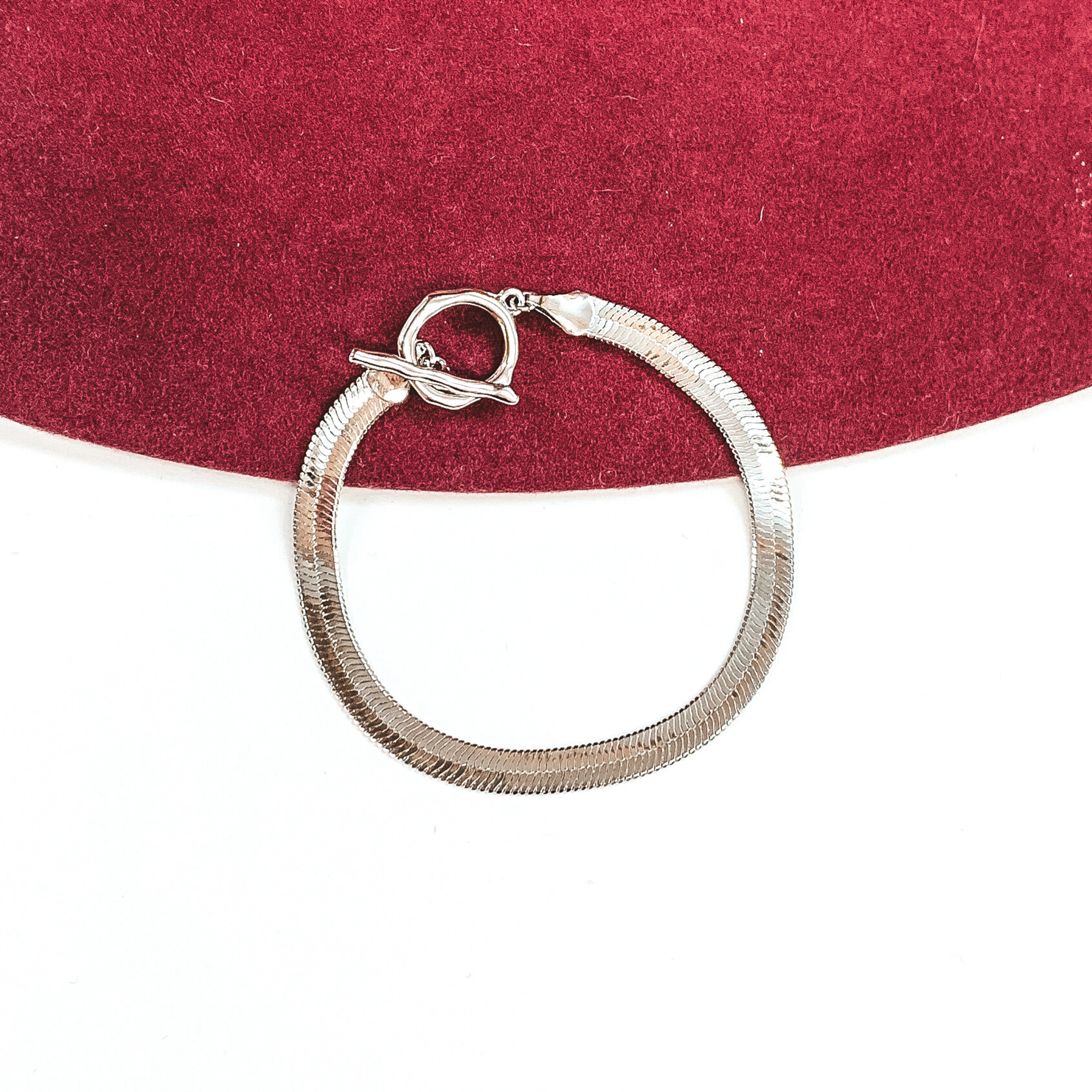 Silver herringbone bracelet that closes with a toggle clasp. This bracelet is pictured on a white and red background. 
