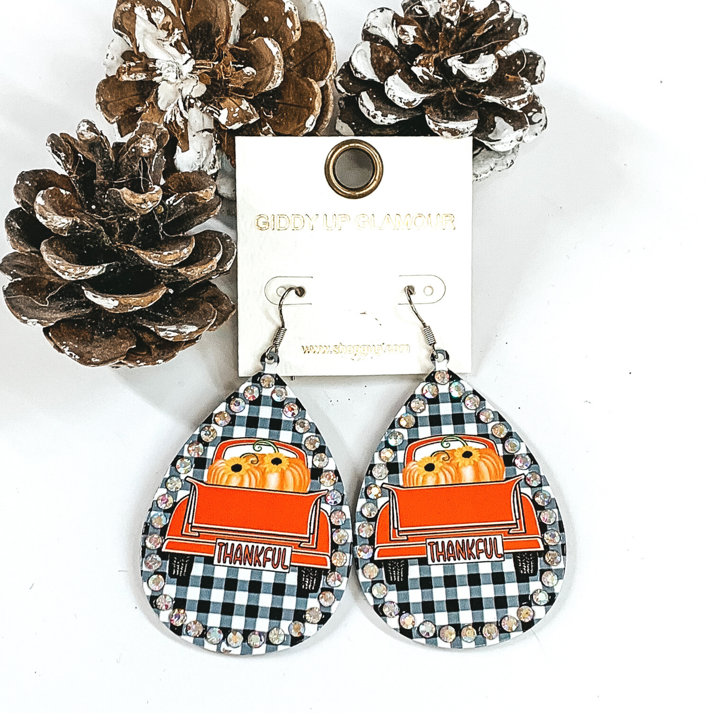 White buffalo plaid tear drop earrings with an ab crystal outline. On the earrings you also have an image of an orange truck bed with orange pumpkins in the bed. These earrings are pictured in a white background with pinecones. 