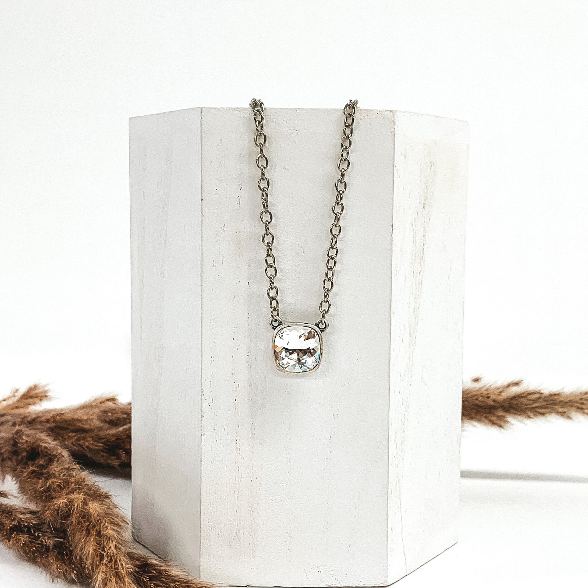 Silver chained necklace with a square clear crystal. This necklace is hanging on a white block next to brown floral decor. 