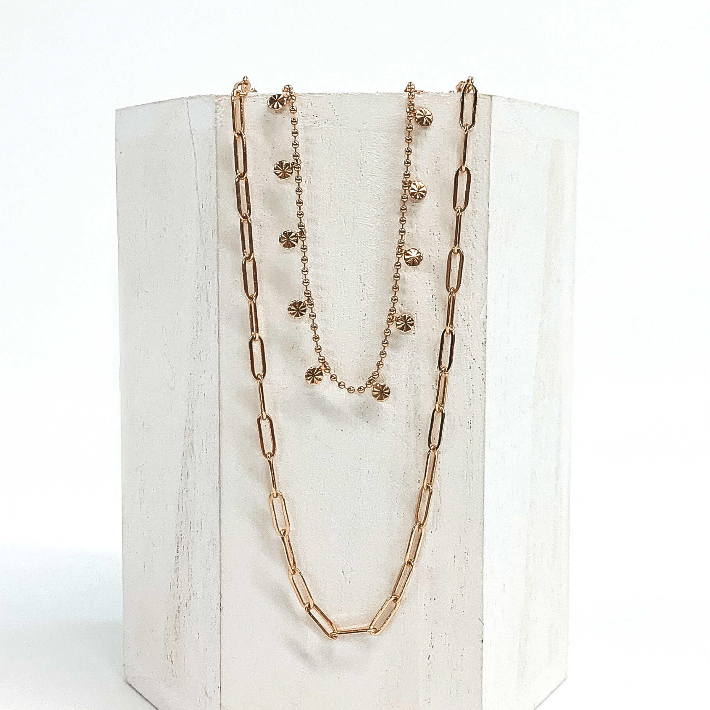 This is a gold double layered necklace. One strand is a thin paperclip chain and the second chain is a beaded chain with tiny charms with a sunburst design. This necklace is pictured on a white block and on a white background.