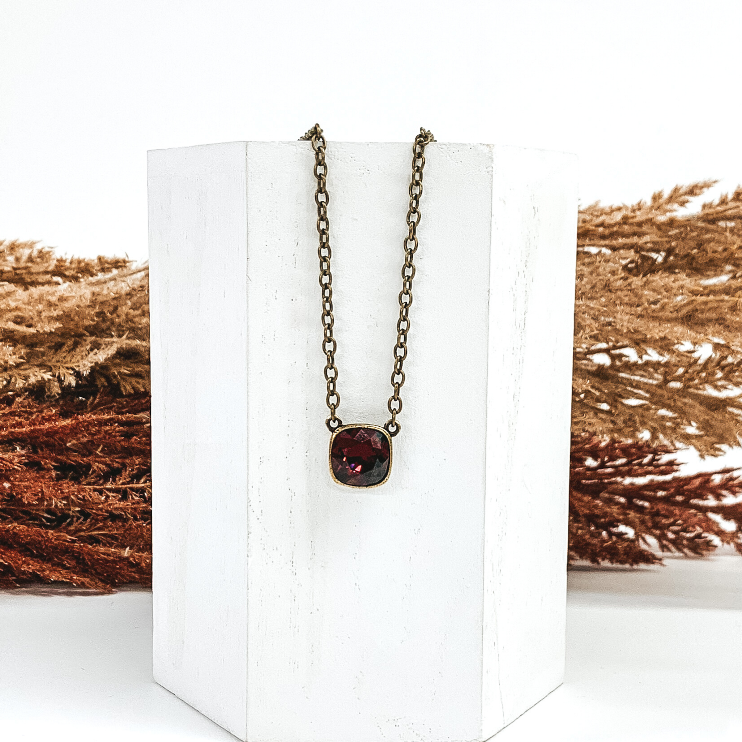 Bronze chained necklace witha square maroon crystal. This necklace is hanging on a white block in front of brown and tan decor
