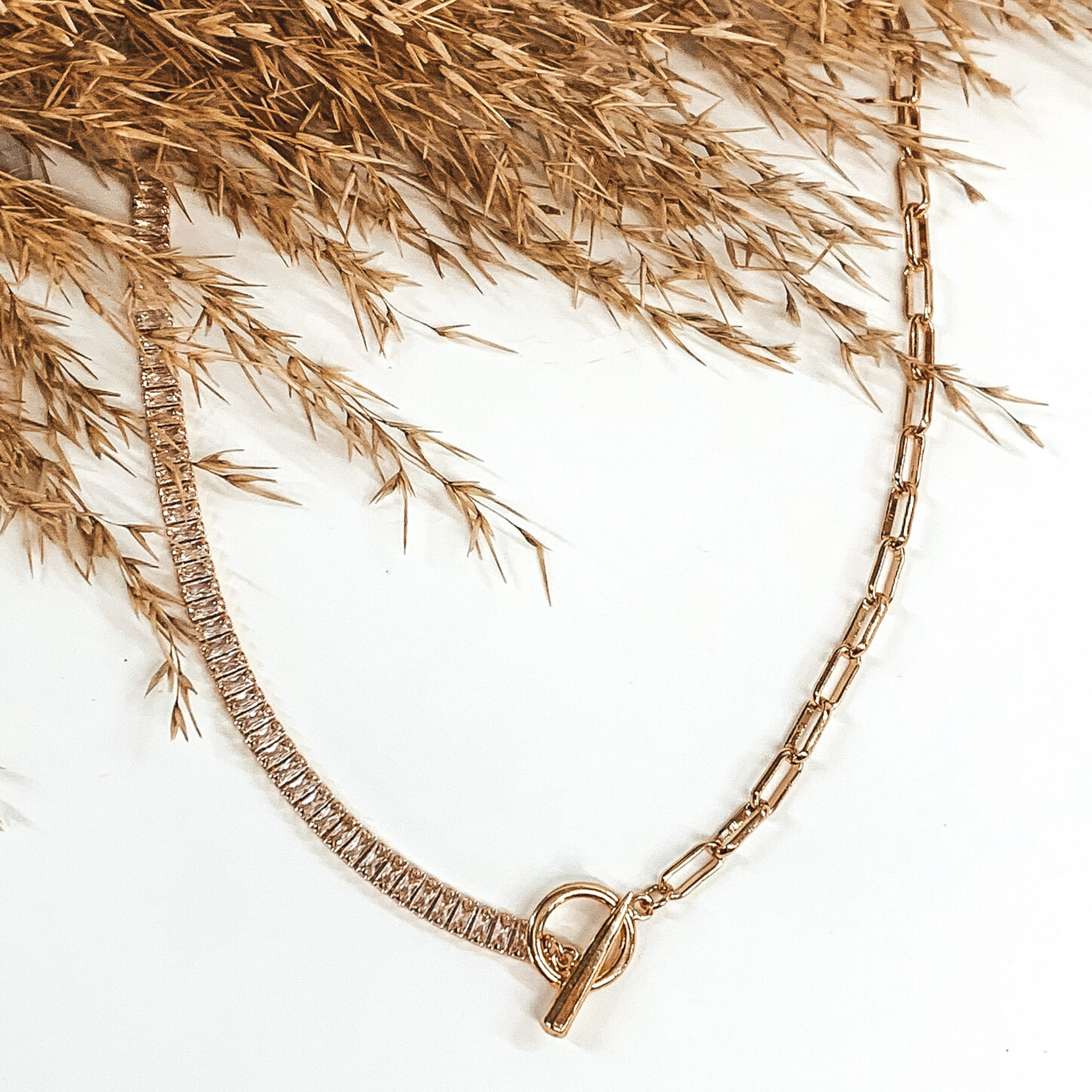 This gold necklace is half clear rhinestones and half thing paperclip chain. There is also a front toggle clasp. This necklace is pictured on a white background with tan floral at the top of the picture.