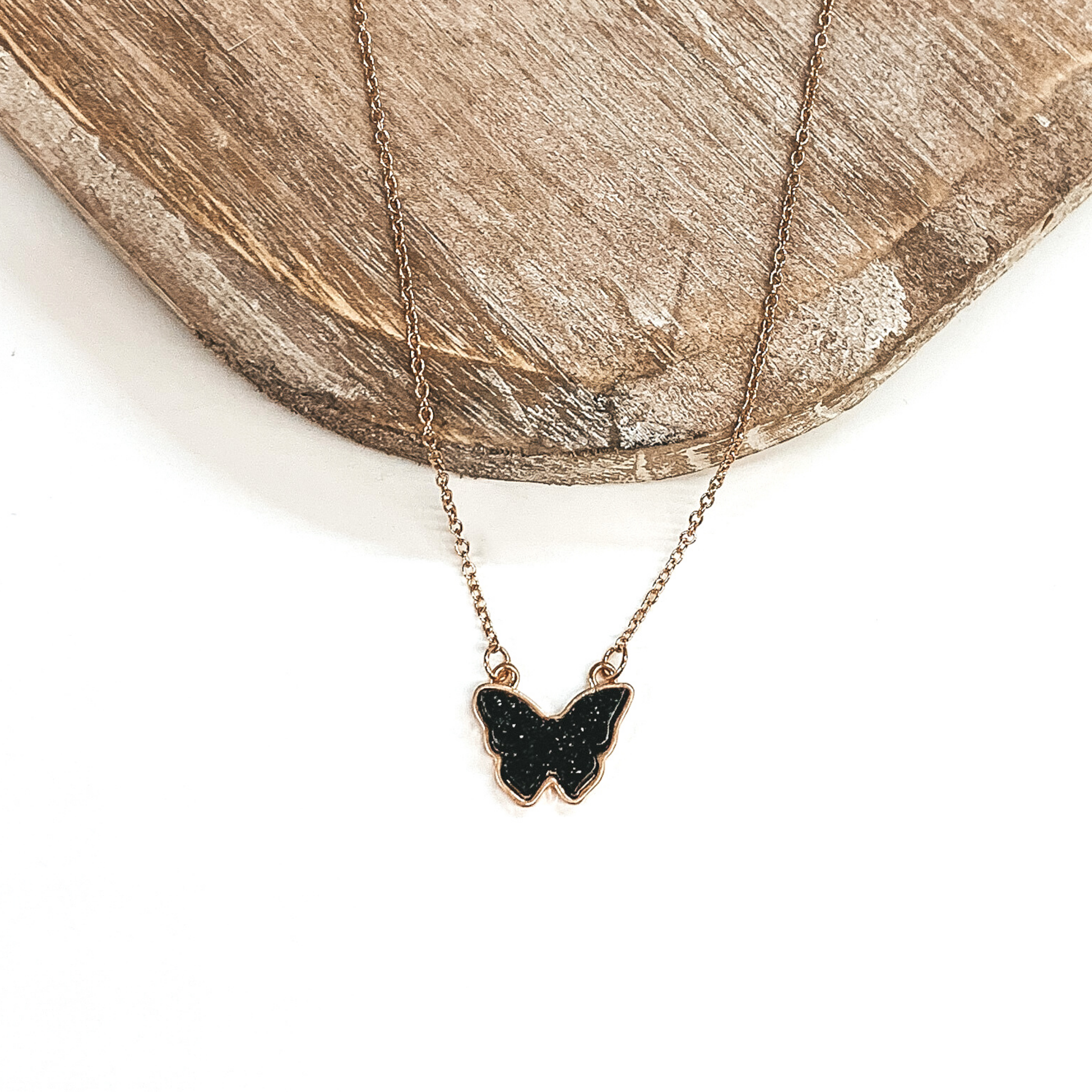 Gold necklace with black druzy butterfly pendant. Pictured on  a white and brown background. 