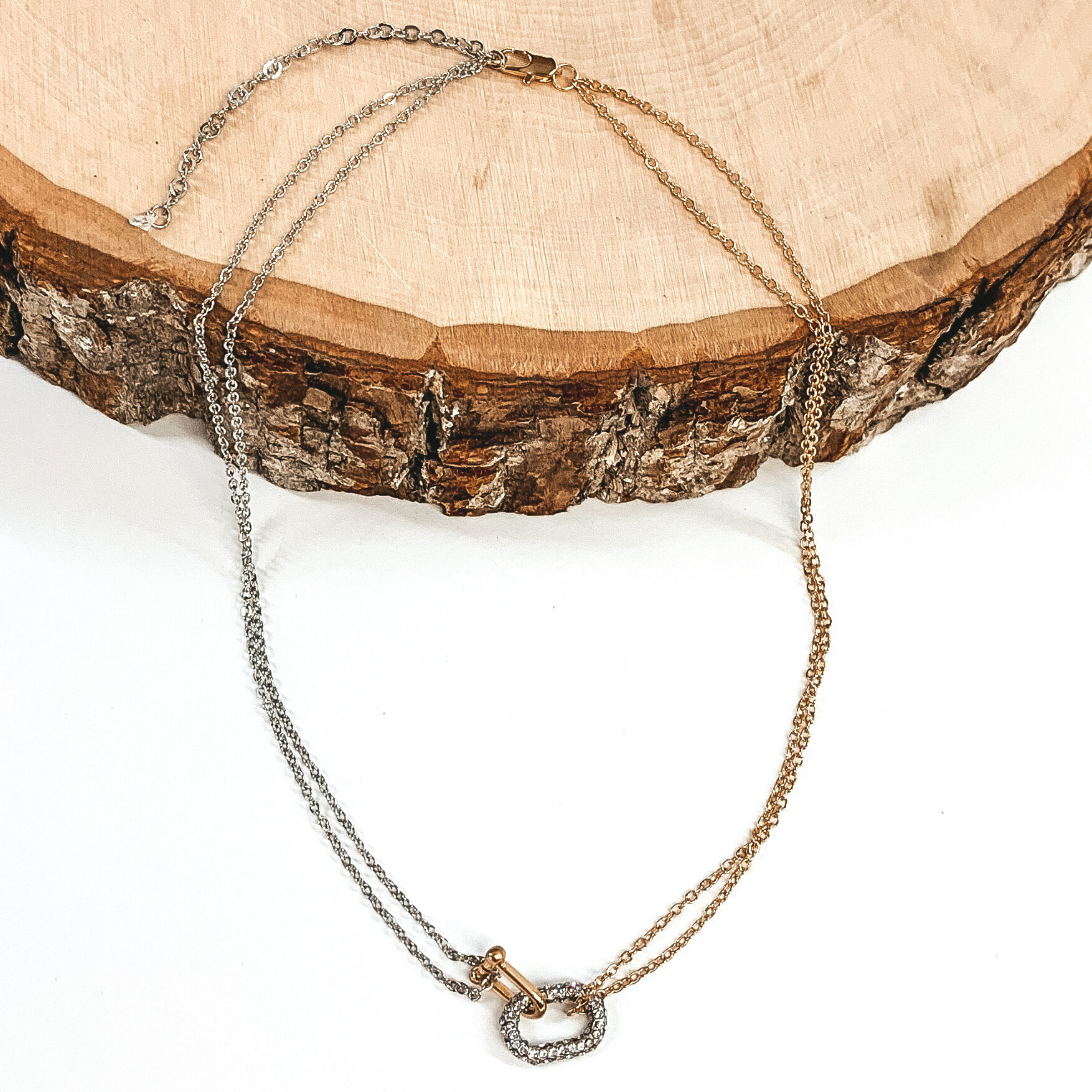 This is a thin, double chained necklace. It is half gold and half silver with a crystal, silver oval pendant and a gold horse shoe pendant. This necklace is pictures on a white background, laying half on a piece of wood. 
