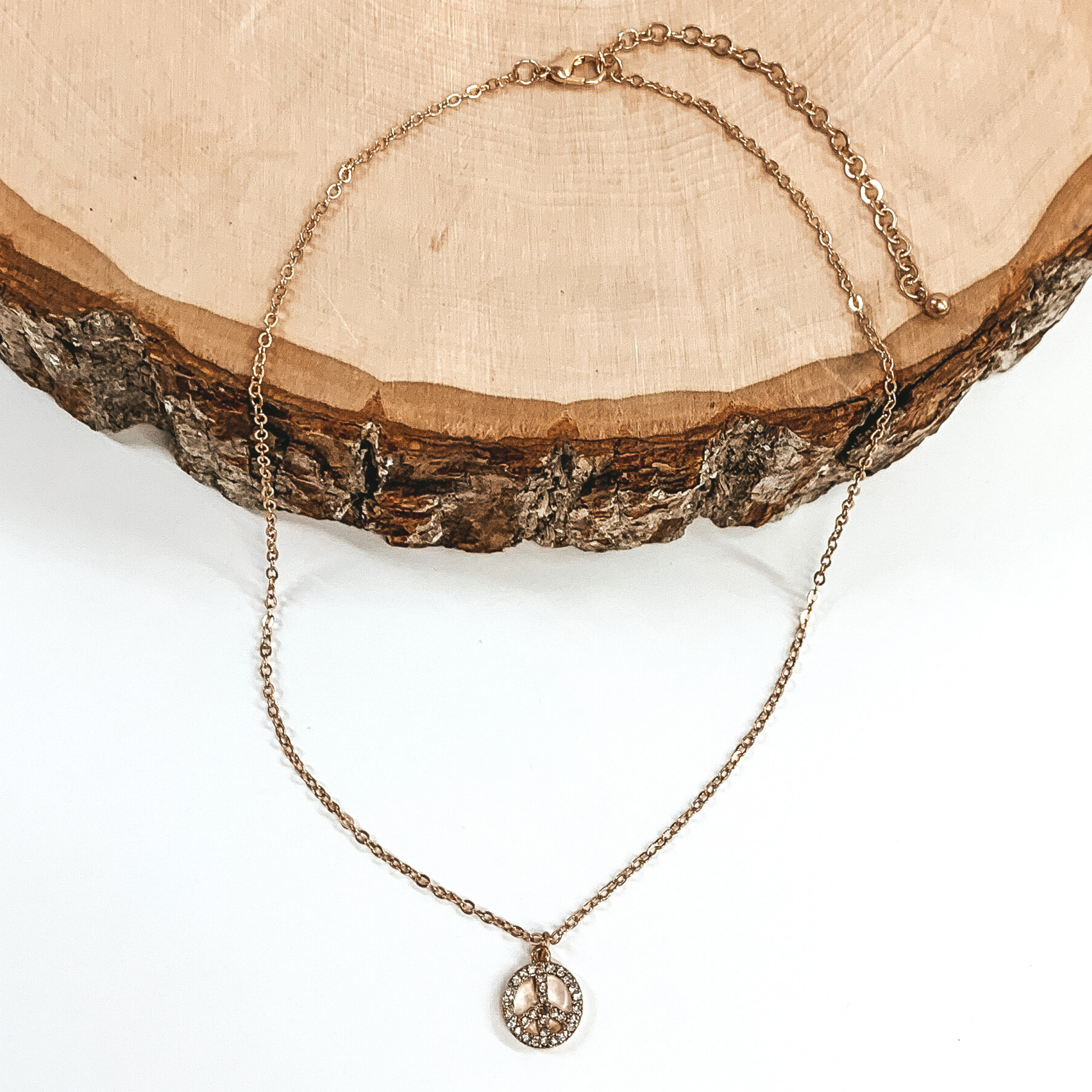 This is a gold thin chained necklace with a small gold, peace sign pendant that has clear crystals. This necklace is pictured half laying on a piece of wood and on a white background.